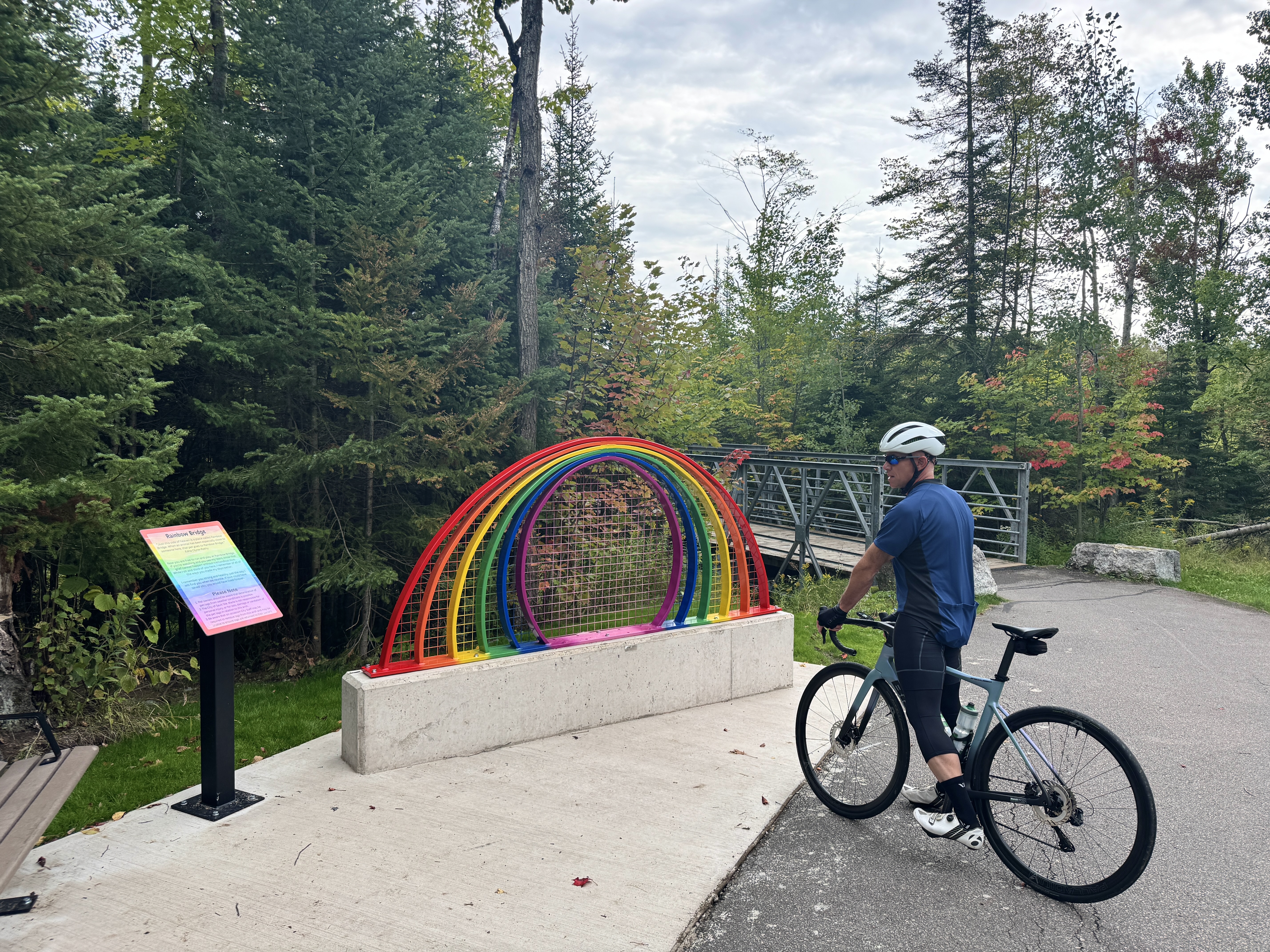 a man on a mountain bike stops to read an informational plaque next to a rainbow statue along a paved bike trail lined with green forest. 