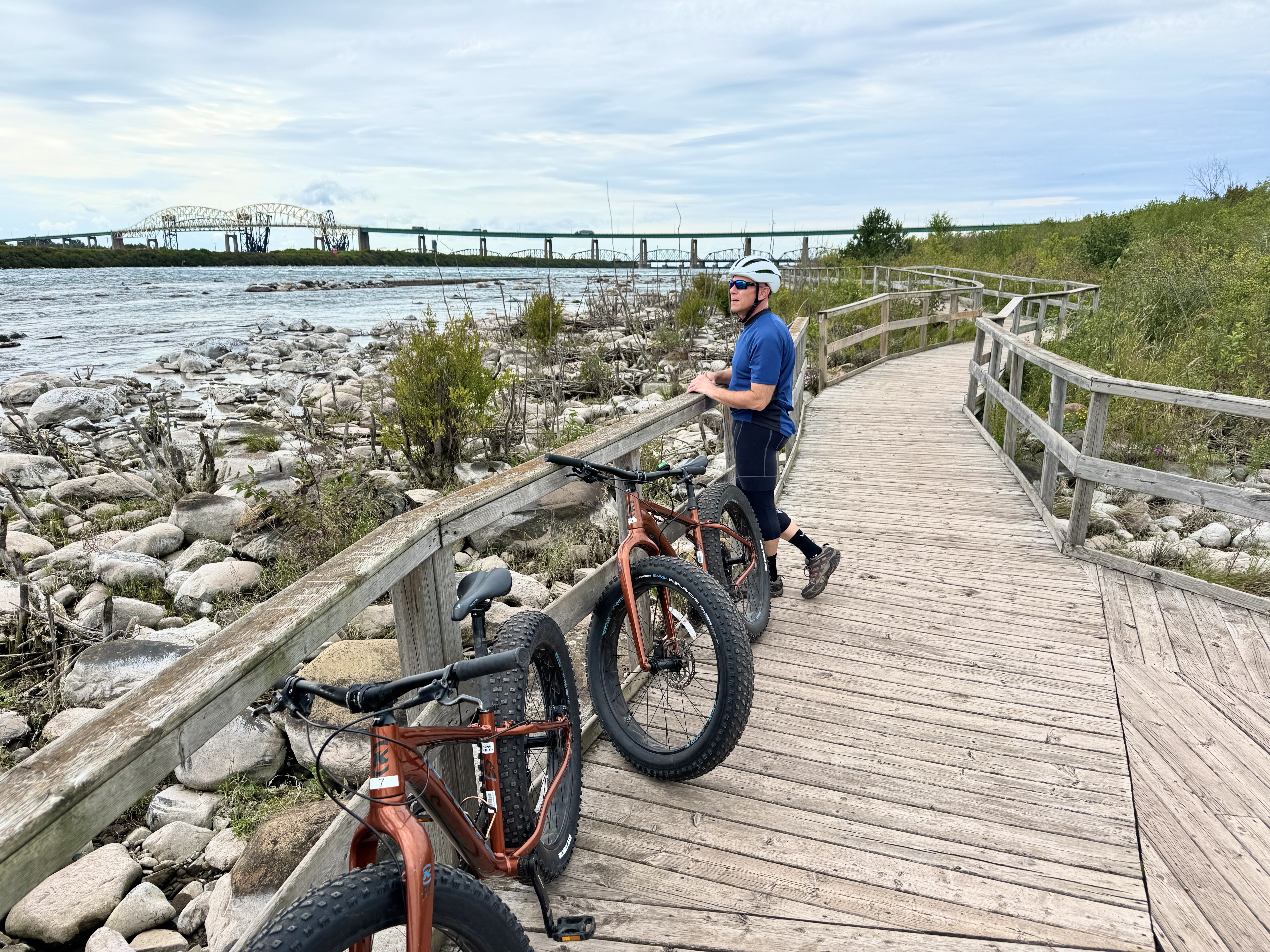 A cyclist stands by two fat bikes stopped on a long wooden boardwalk next to the rocky-shored Sault Canal.