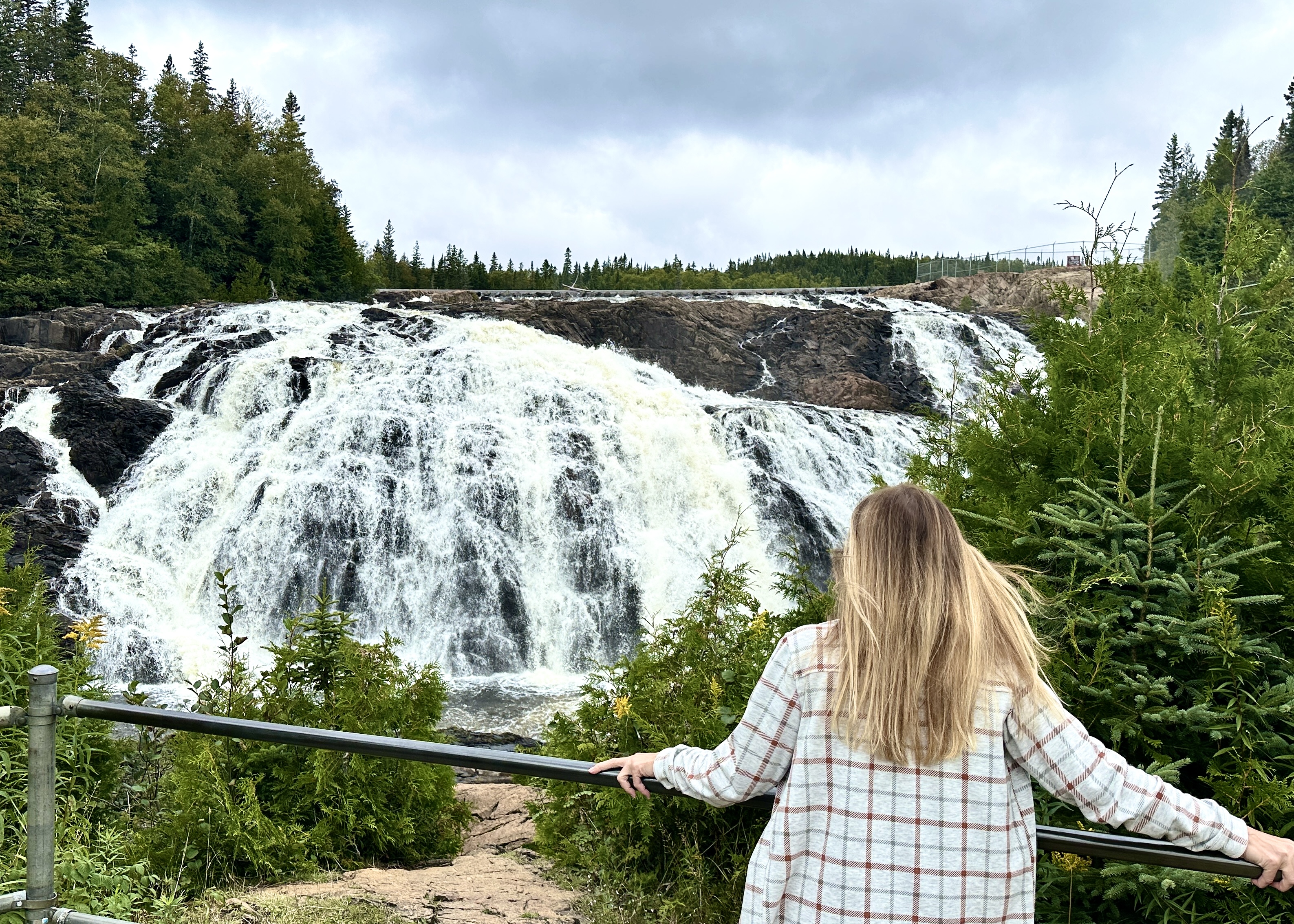 A woman stands at a metal railing, looking out over a tall rushing watefall over a rock cliff surrounded in green forest. 