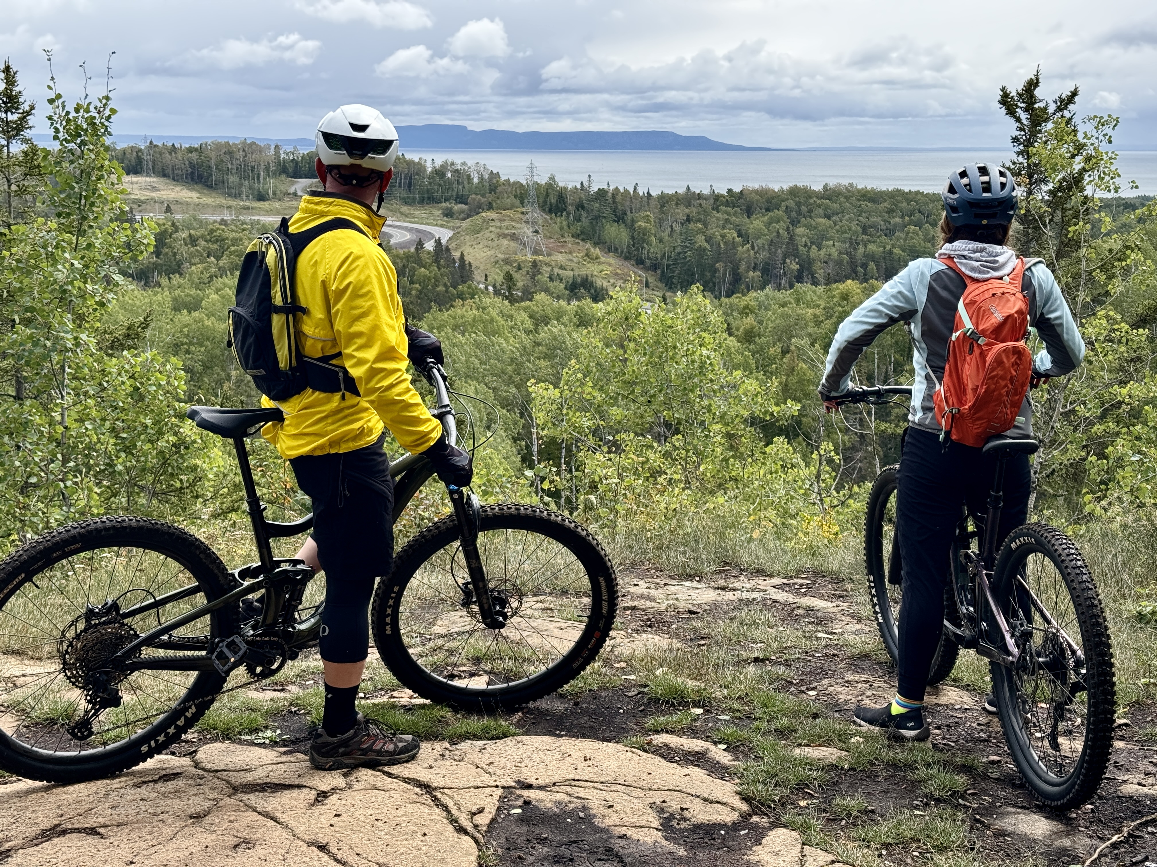 Two people stand with their mountain bikes at a smooth, rocky lookout peering out over a wide green forested valley with Lake Superior and Sleeping Giant on the horizon. 
