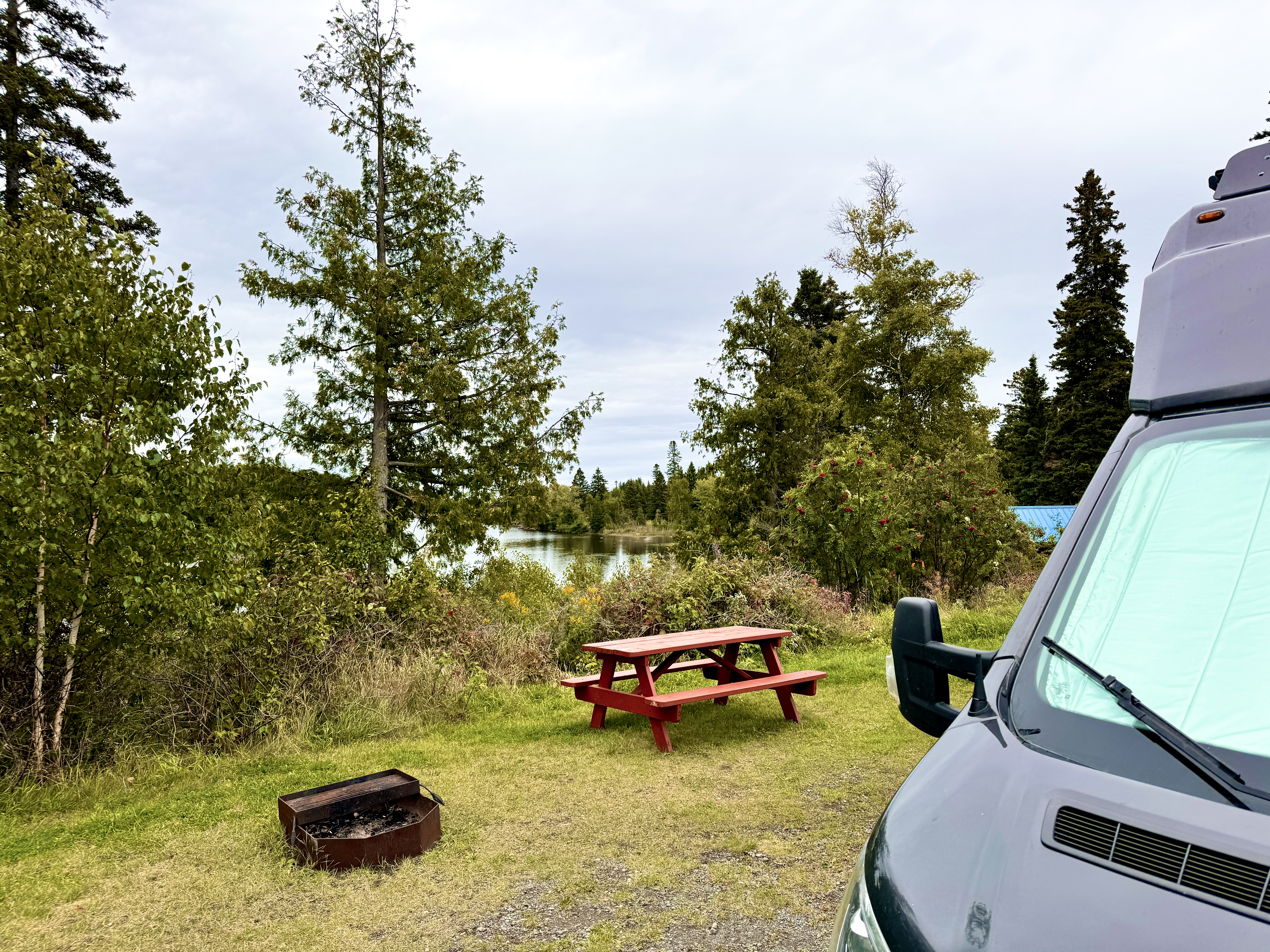 An RV parked at a green, forested lakeside campsite with wooden picnic table and an iron fire ring. 