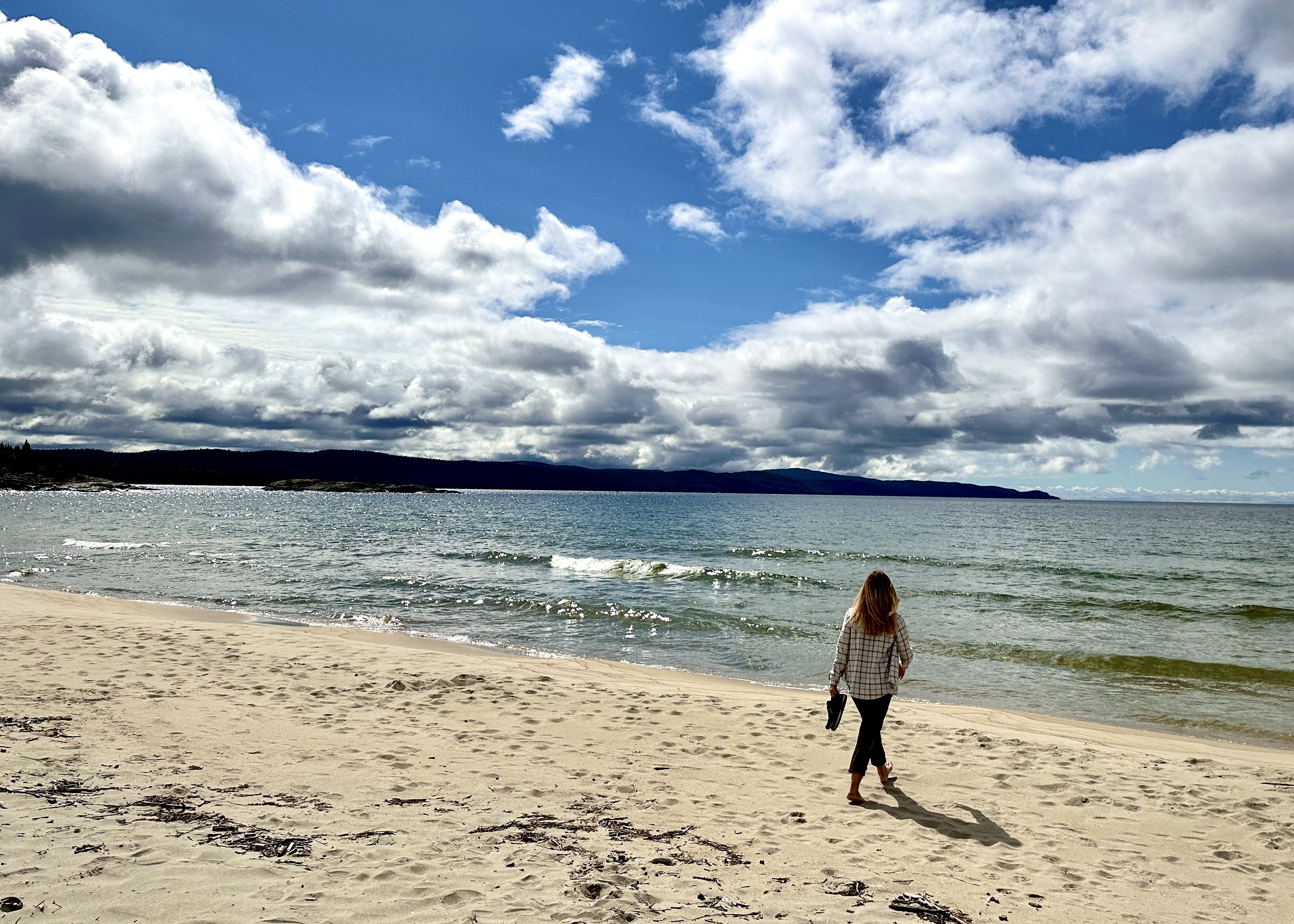 A woman walks with her shoes in hand across a soft white sandy beach, looking out over the sparkling turquoise Lake Superior waves. 