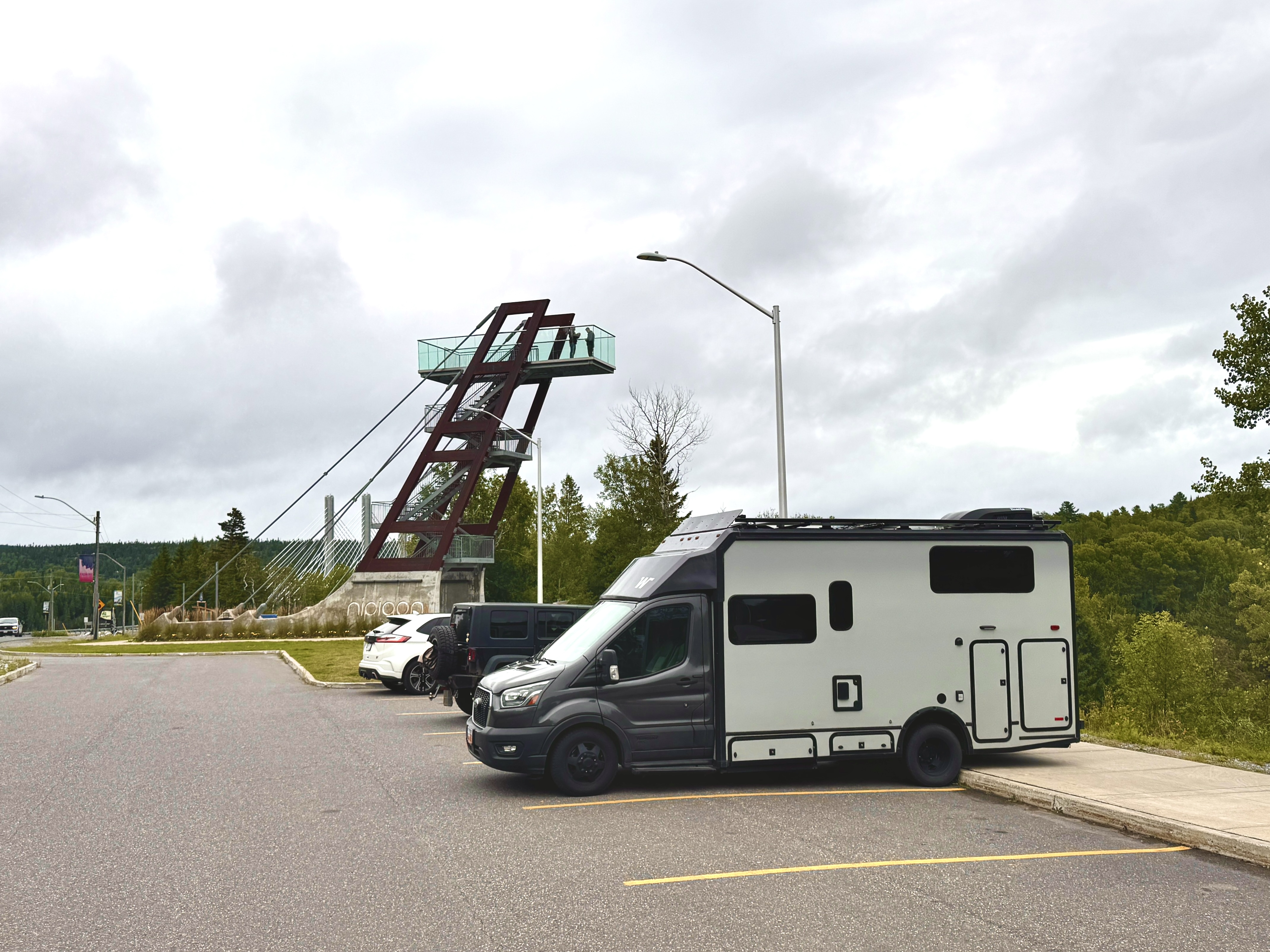 An RV parked next to the Nipigon Lookout, a tall metal structure with stairs leading to a glass-enclosed platform high above the green trees.