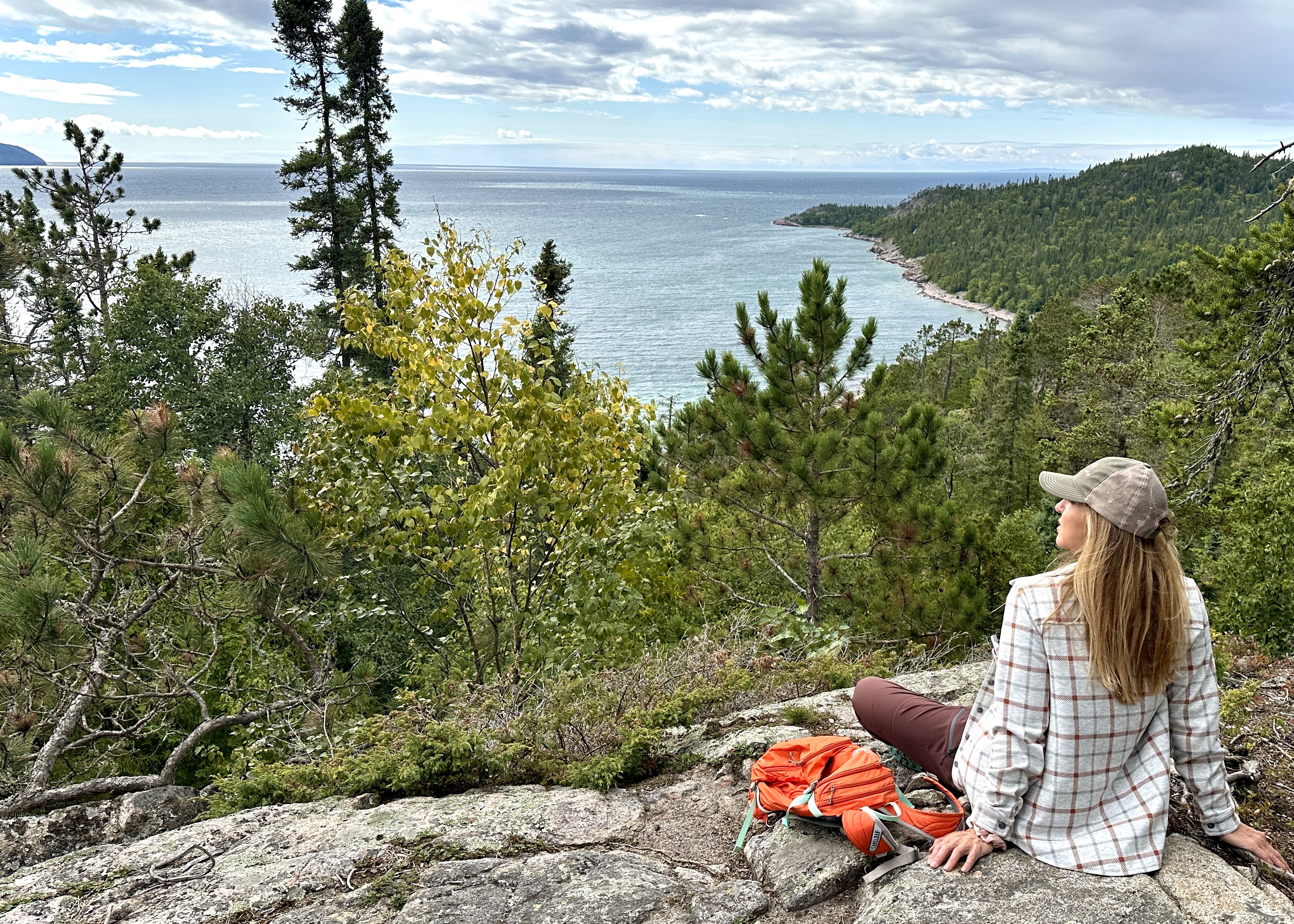 A woman sits on a rock cliff overlooking a green forested valley and the broad, blue Old Woman Bay on Lake Superior.