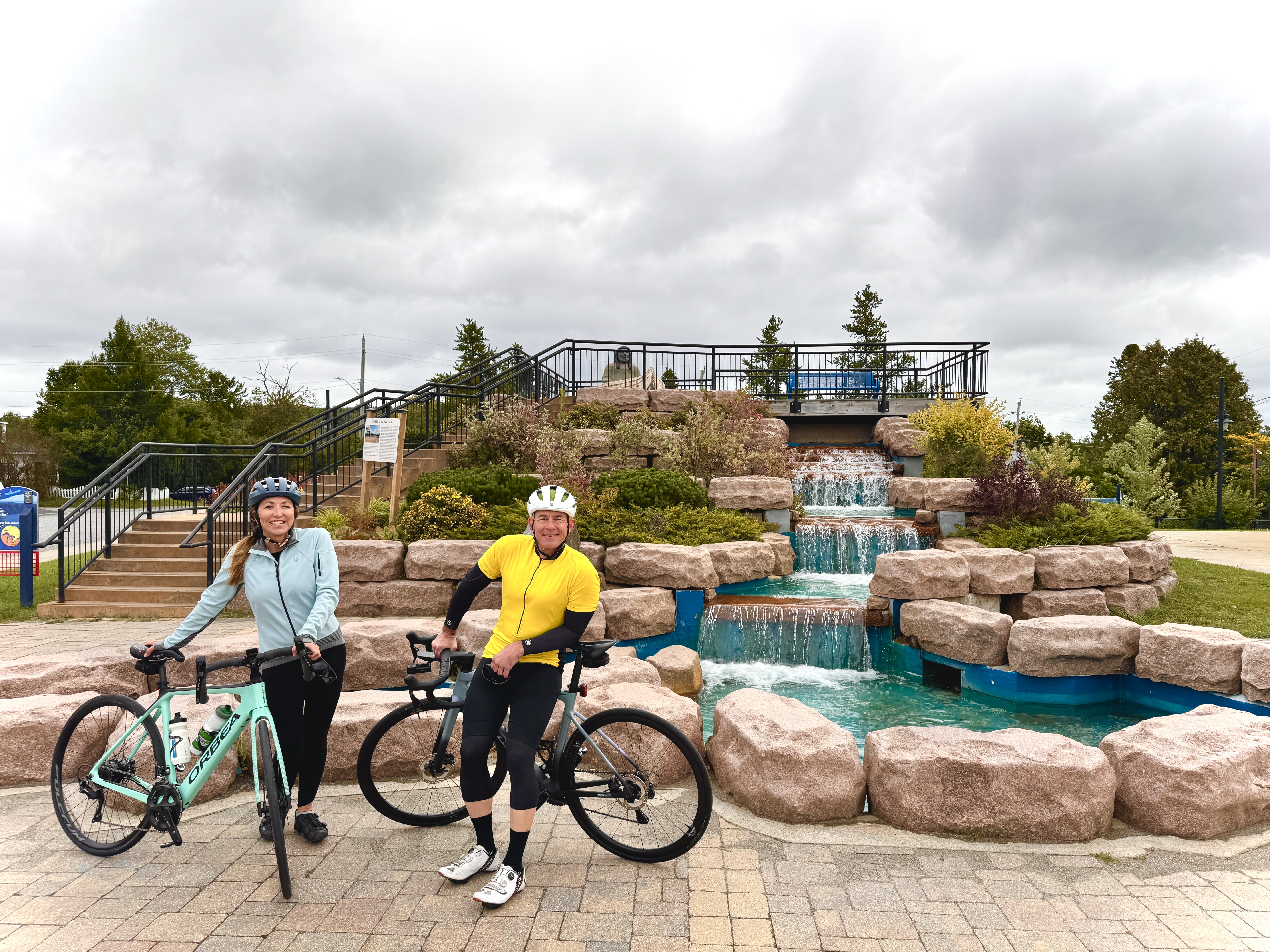 Stef and James of The Fit RV standing next to their mountain bikes in front of a rock waterfall fountain at Paddle to the Sea Park in Nipigon. 