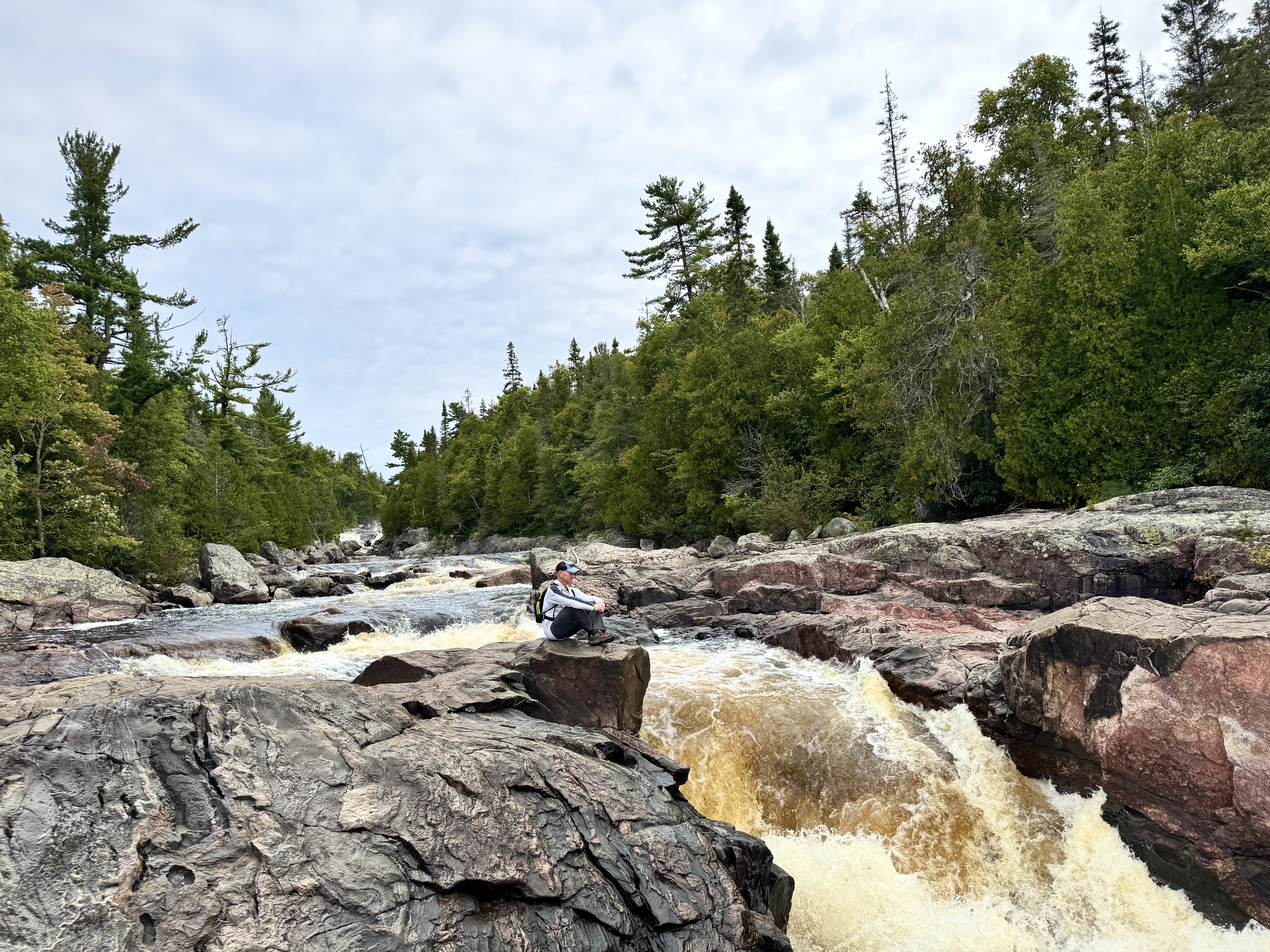A man sits on a rock cliff overlooking a short, rushing waterfall in a rocky riverbed surrounded by thick forest.