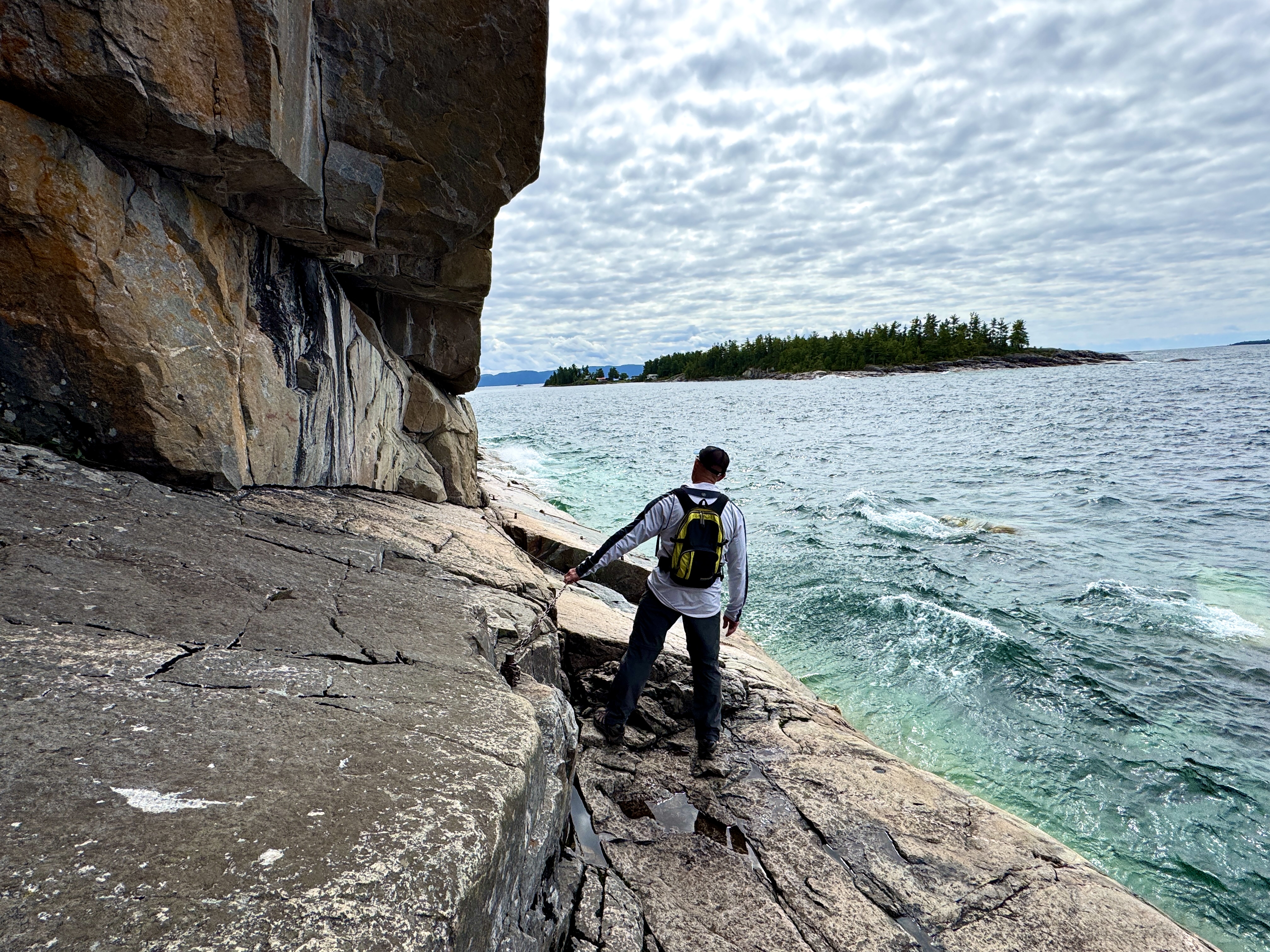 a man wearing a backpack walks along a narrow rock bank skirting a tall cliff overhang next to the deep turquoise water of Lake Superior.