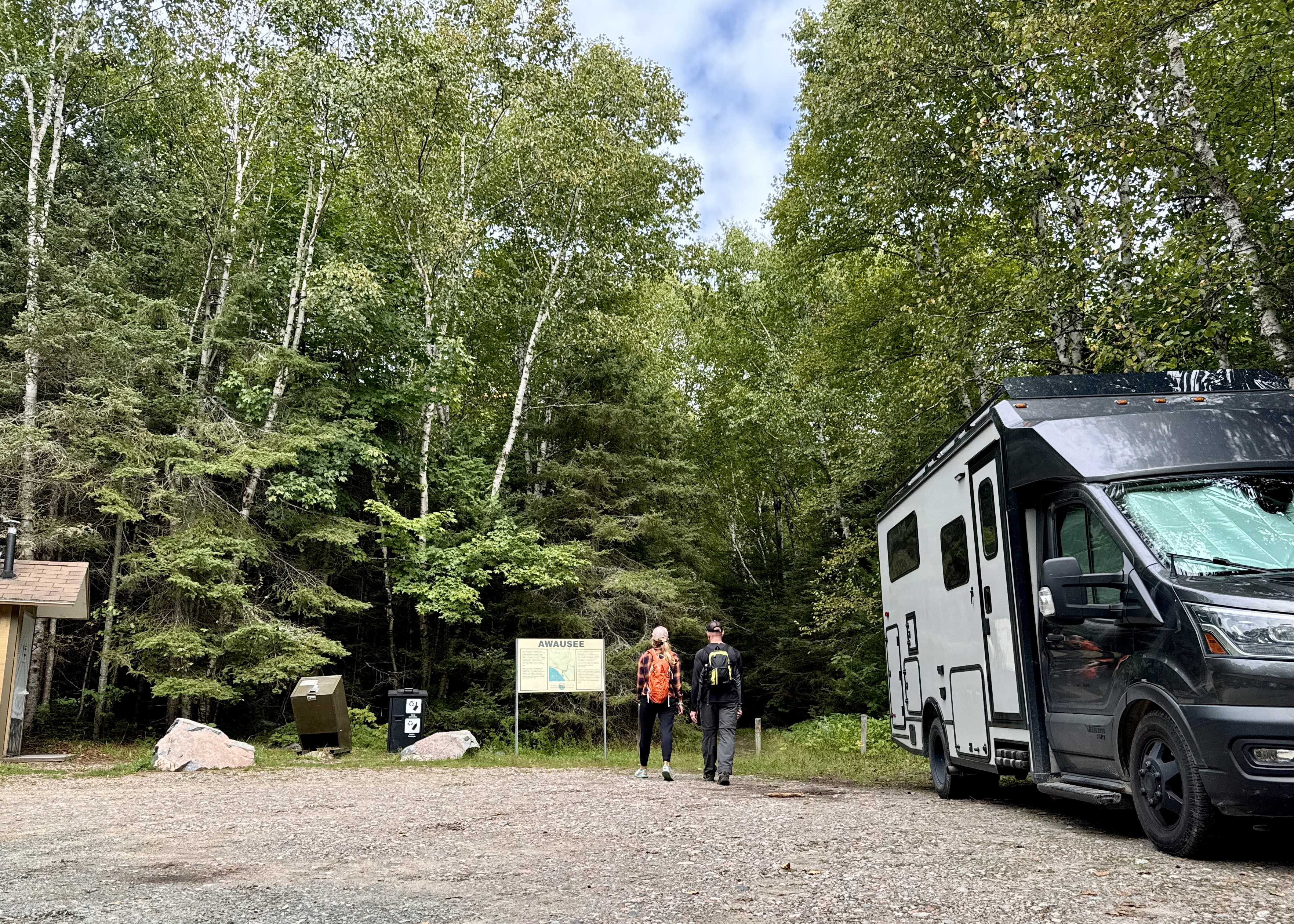 A man and woman wearing backpacks walk to the Awausee Trail head from their RV parked next to thick green forest. 