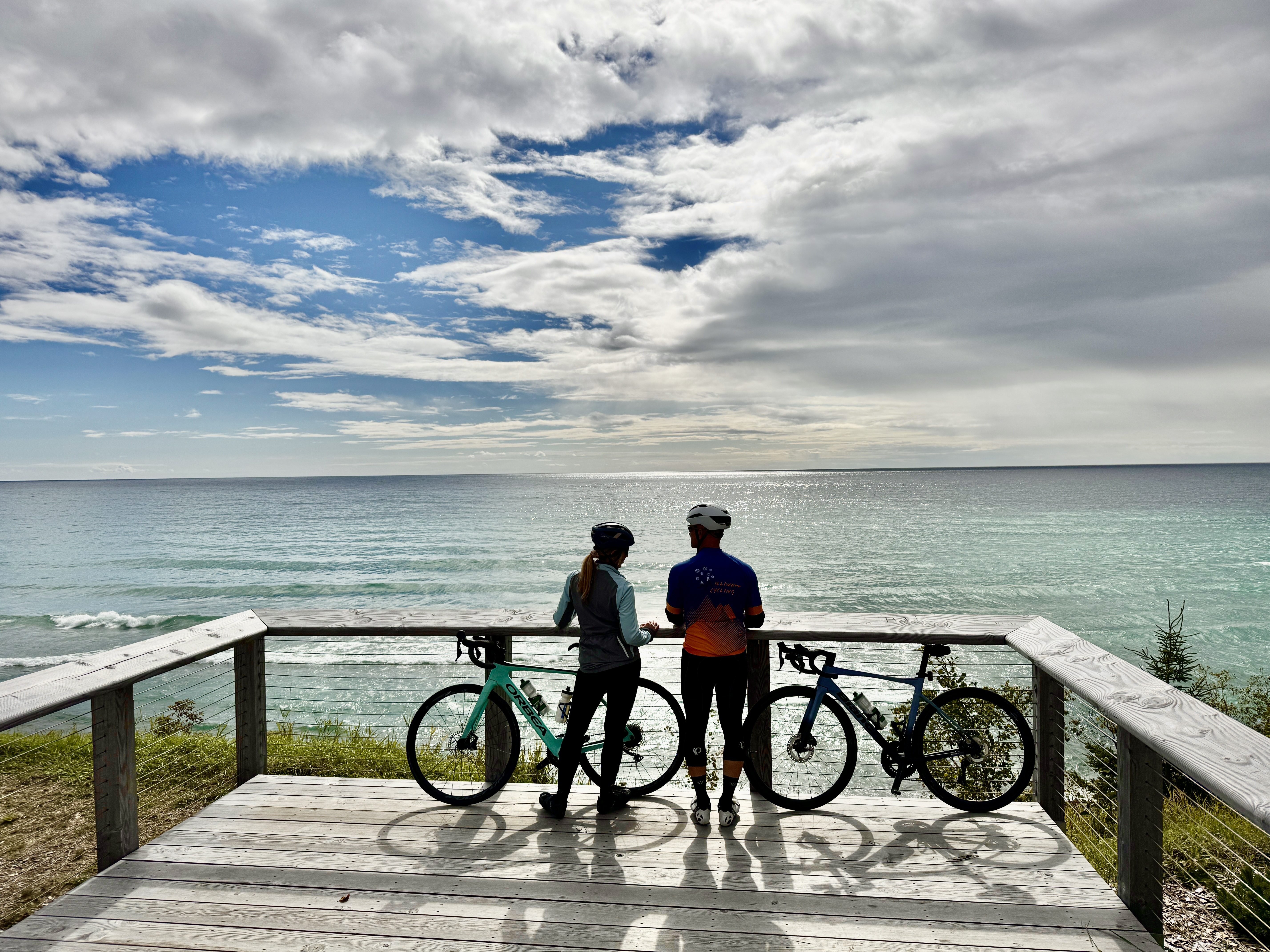 a man and woman stand with their mountain bikes on a wooden platform looking out over Pebble Beach while the sun gleams off the vast turquoise water of Lake Superior.