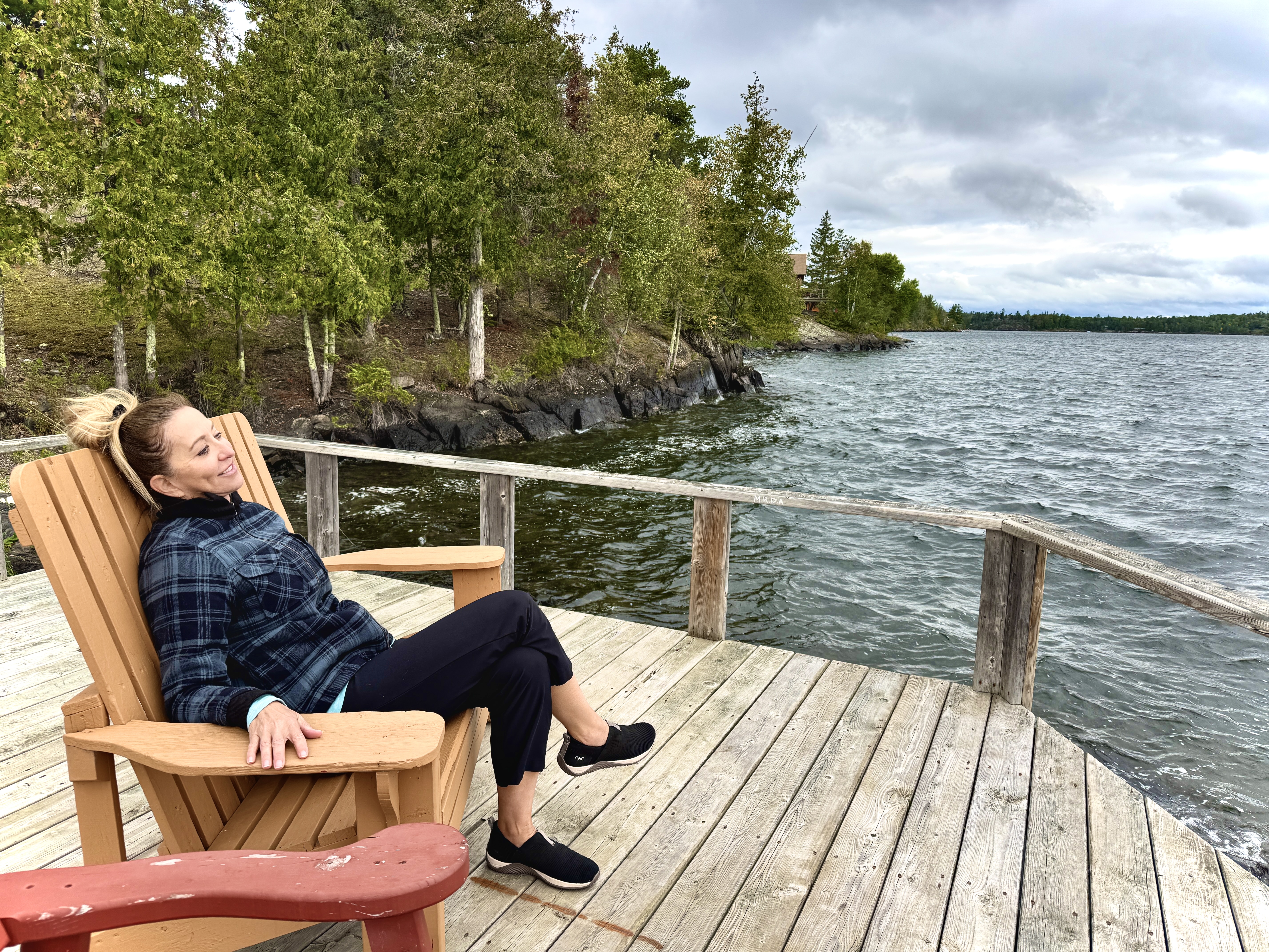 A woman smiles as she lounges in a wooden chair on a deck overlooking a forest lake at Tomahawk Resort in Sunset Country. 