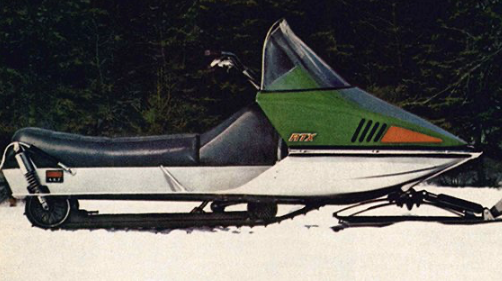 A green and orange 1972 Skiroule RTX 440, a trim, pointed and streamlined snowmobile parked in front of a pine forest. 