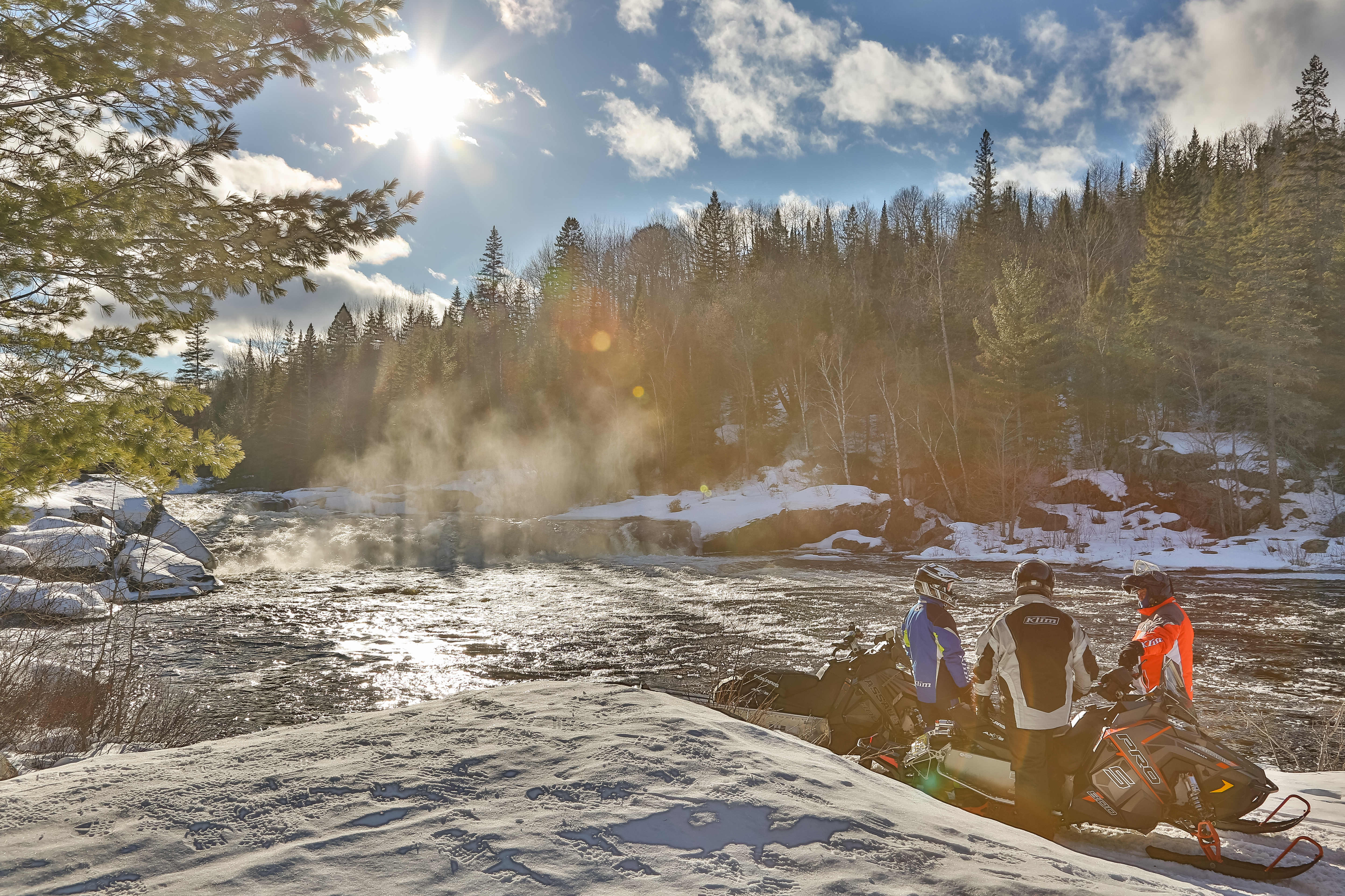 two snowmobilers chat next to their machines in a snowy forested valley lit up by a golden winter sunset by Lake Kukagami.