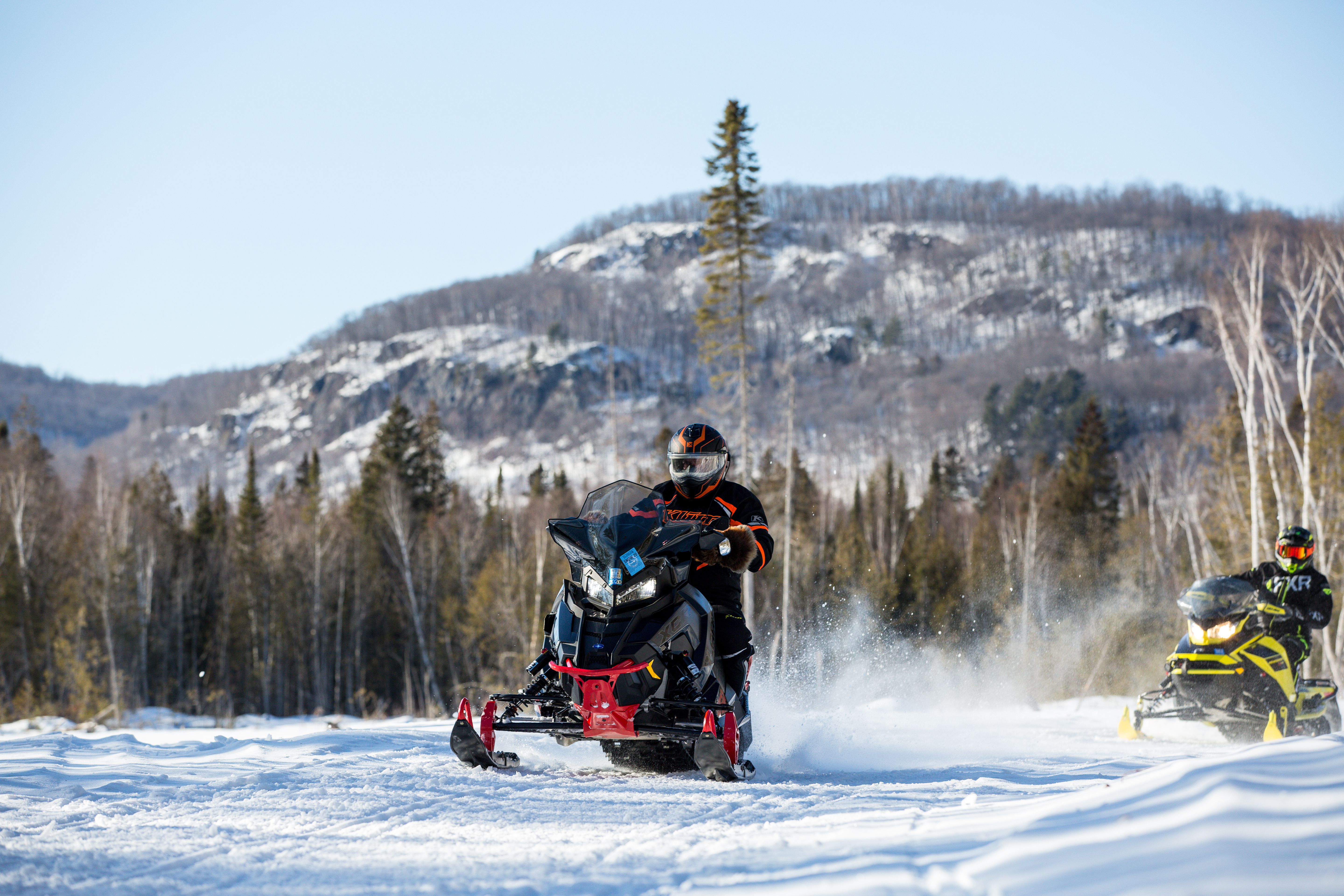 a snowmobiler riding along a powdery ridge near Sudbury, Ontario with tall forested hills in the background.
