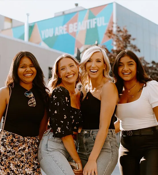 a group of female friends smile, laugh and stand close together under a mural in Sudbury that reads "You Are Beautiful" on a sunny summer day.