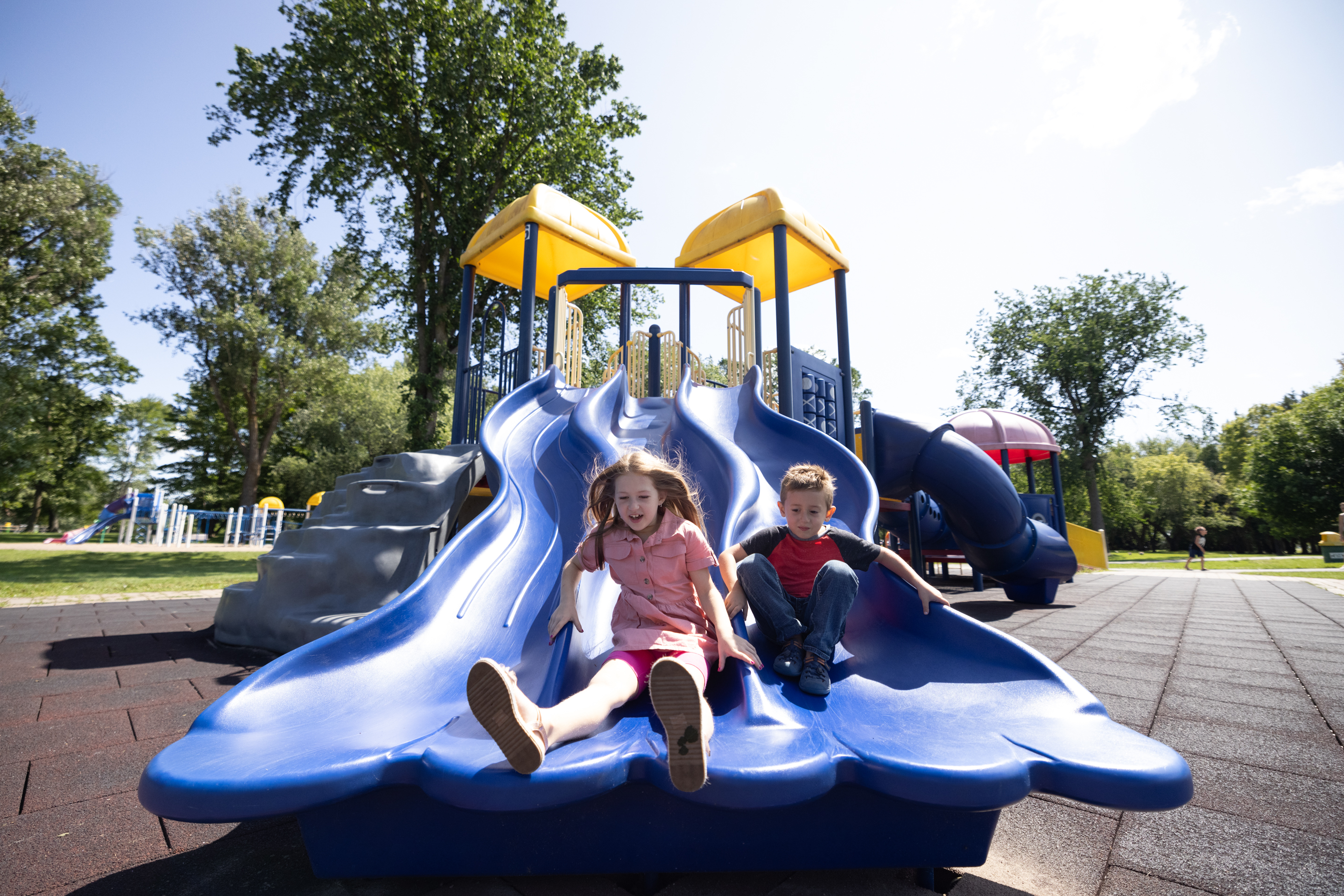 Children laugh as they slide down a tall, winding plastic slide on a sunny summer day at Bellvue Park.