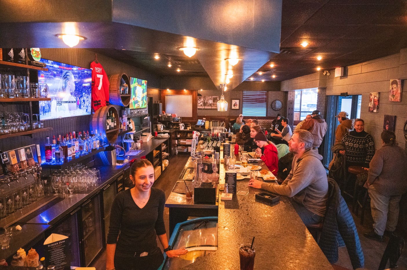 a smiling waitress greets patrons while several people sit watching a sports game at the homey, warmly-lit bar at Burger Don.