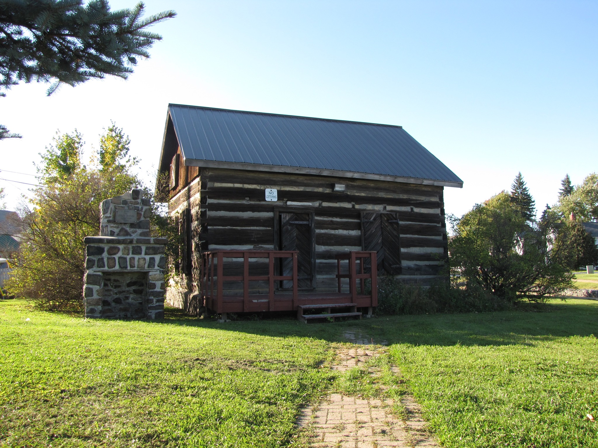 The Copper Cliff Museum, a small log house on a lush green lawn under a bright blue morning sky. 