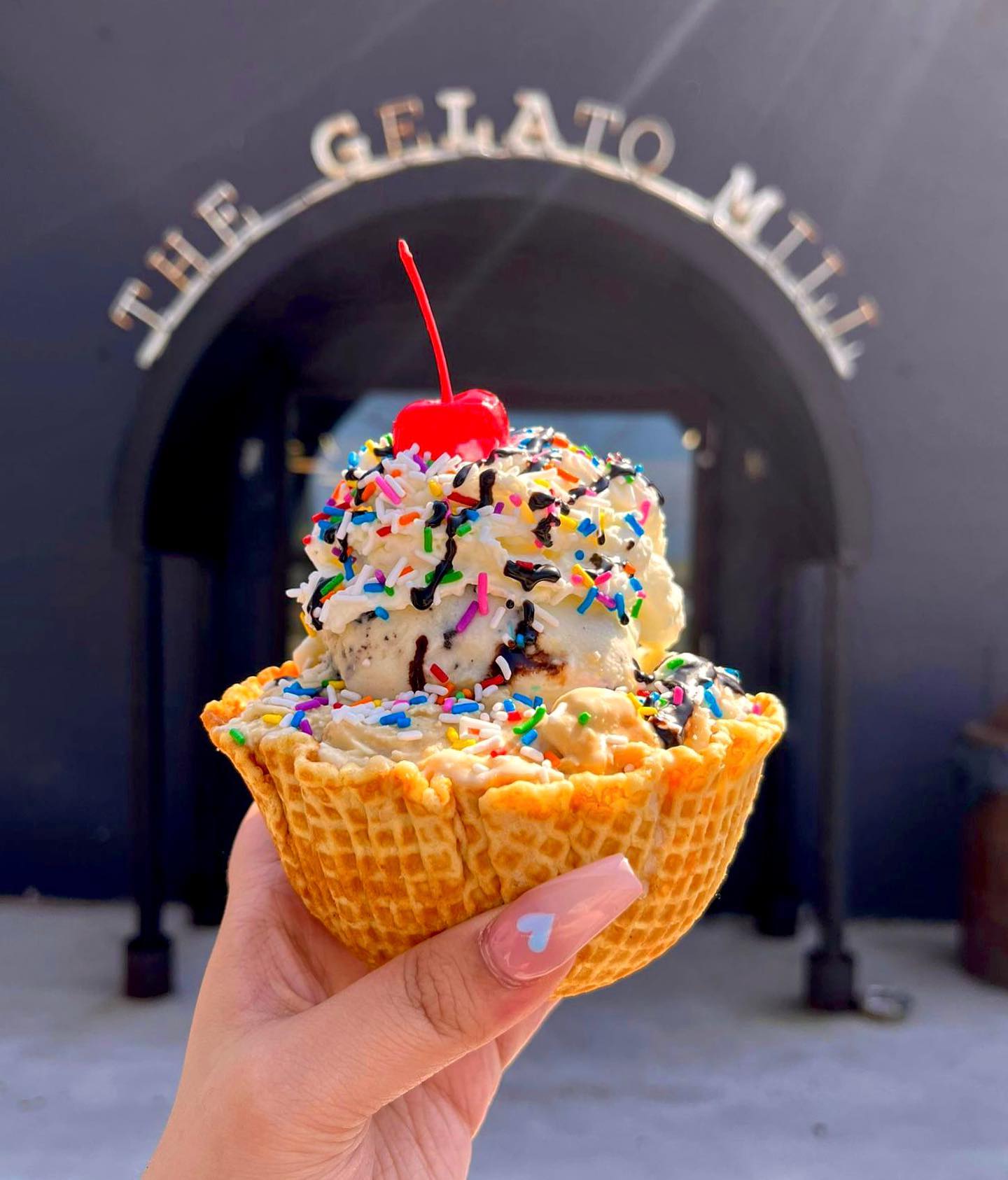 a hand holding a waffle bowl filled with ice cream topped with colourful sprinkles and a cherry in front of a sign that reads " The Gelato Mill".