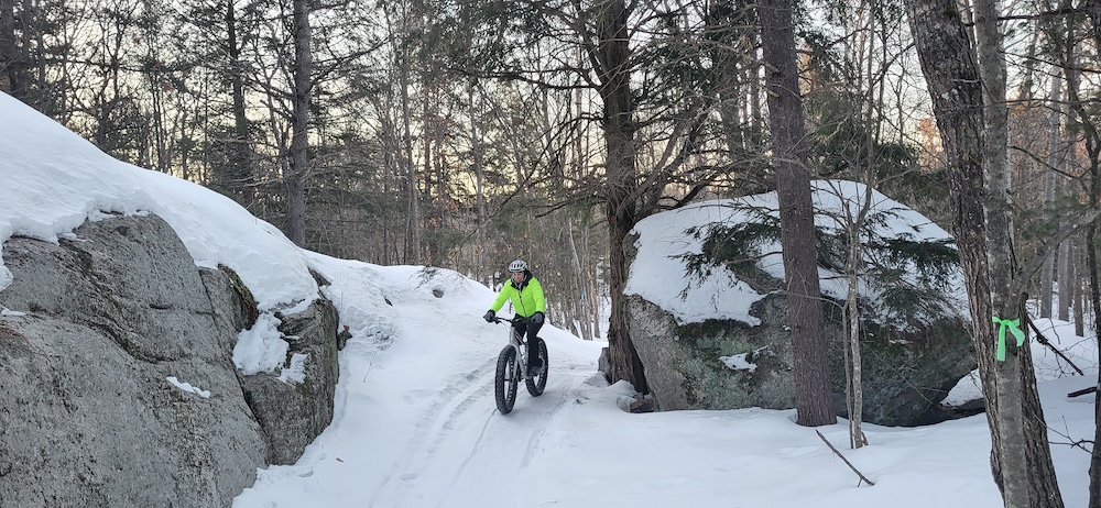 Woman on fat bike going down snowy hill beside two boulders