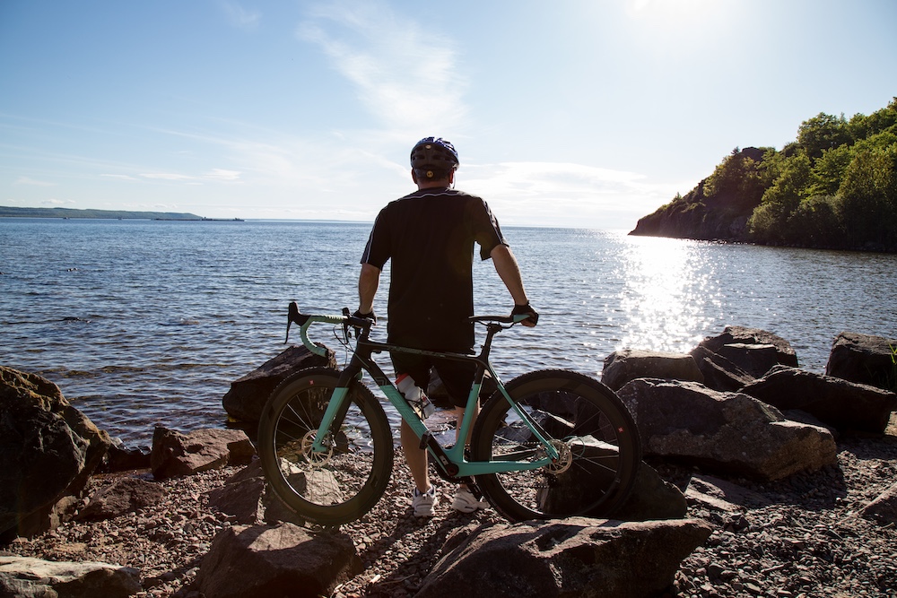 Man stands next to road bike on rocky beach looking out at Lake Superior
