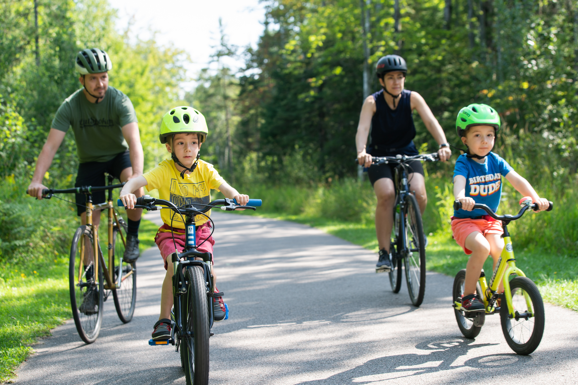 a family rides their bikes down the paved forest trails of the John Rowswell Hub Trail in Sault Ste. Marie on a summer day.