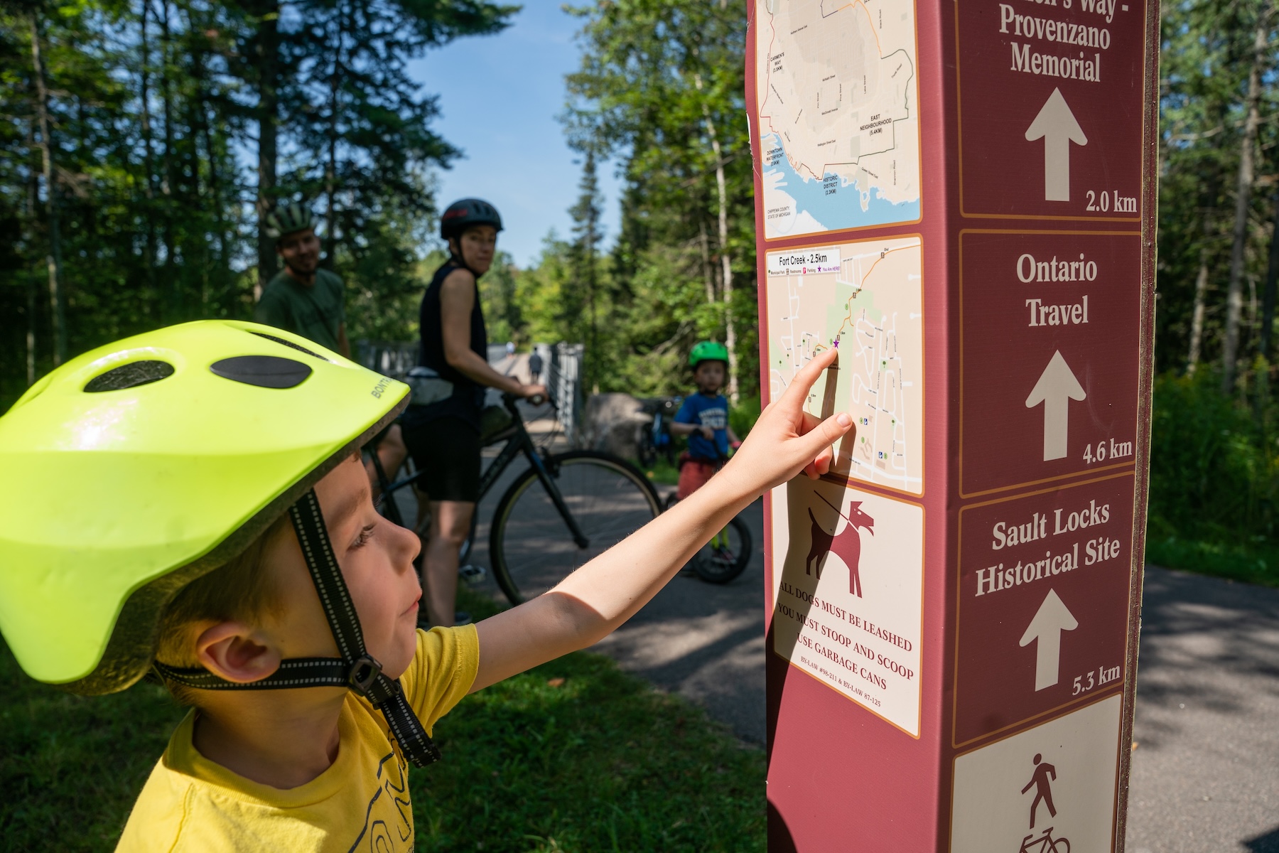 a child in a bike helmet looks at the maps posted on directional marker signs along the John Rowswell Hub Trail.