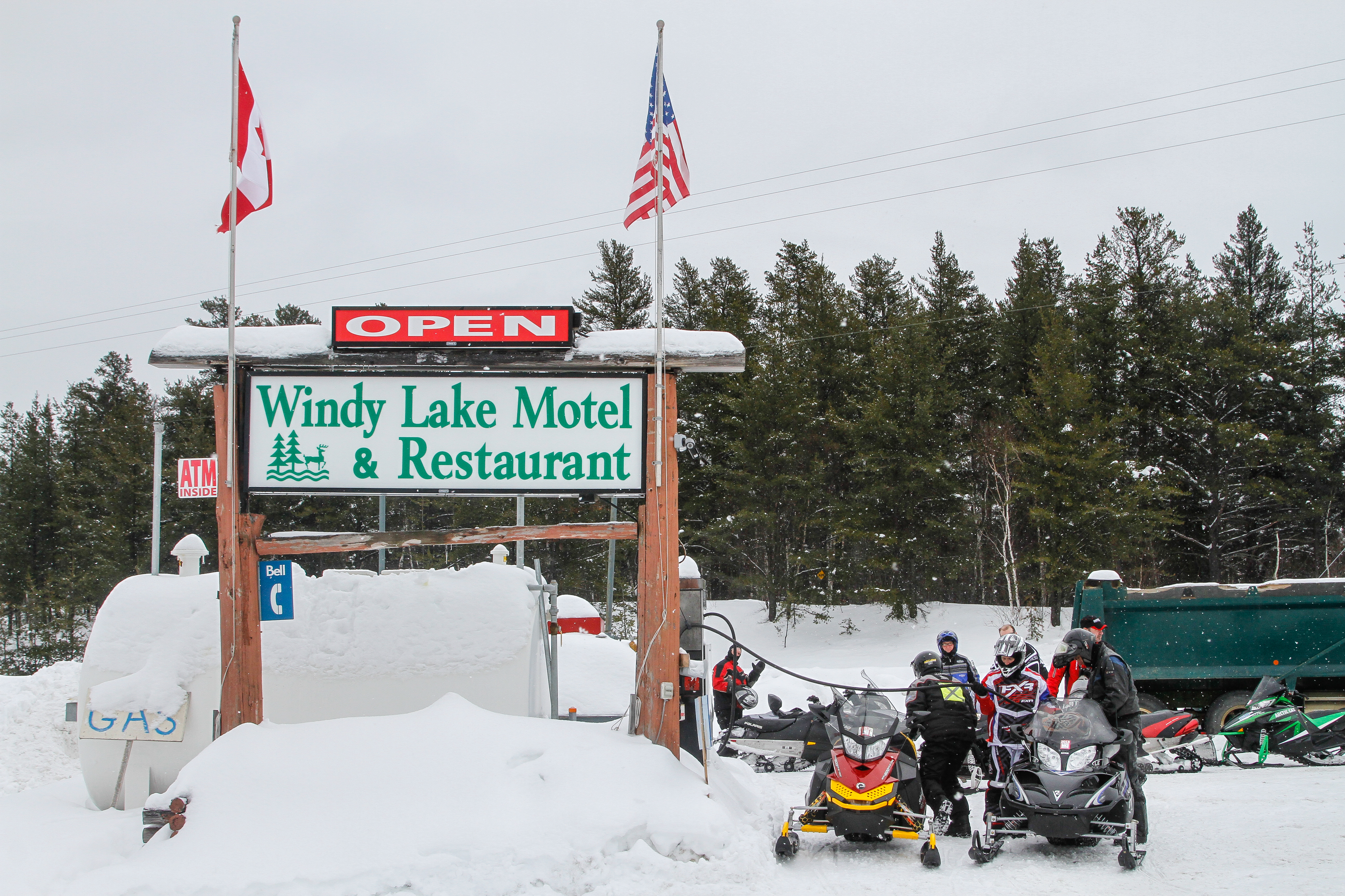 Snowmobilers stand next to their machines by a large, wooden, snow-covered sign for the Windy Lake Motel and Restaurant.  