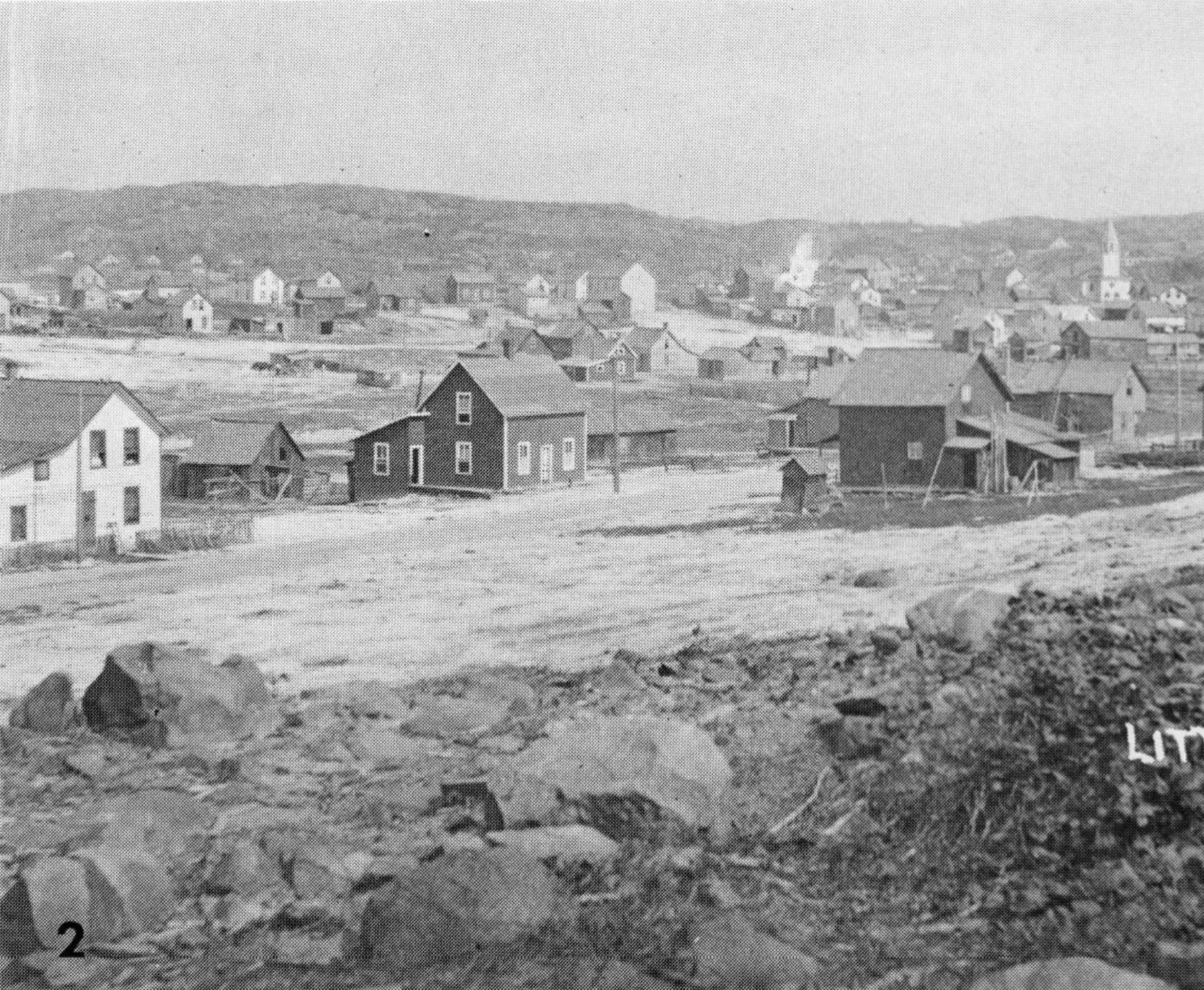 A black and white photograph of Little Italy in Copper Cliff in 1912, with small wooden buildings dotting a flat, rocky landscape. 