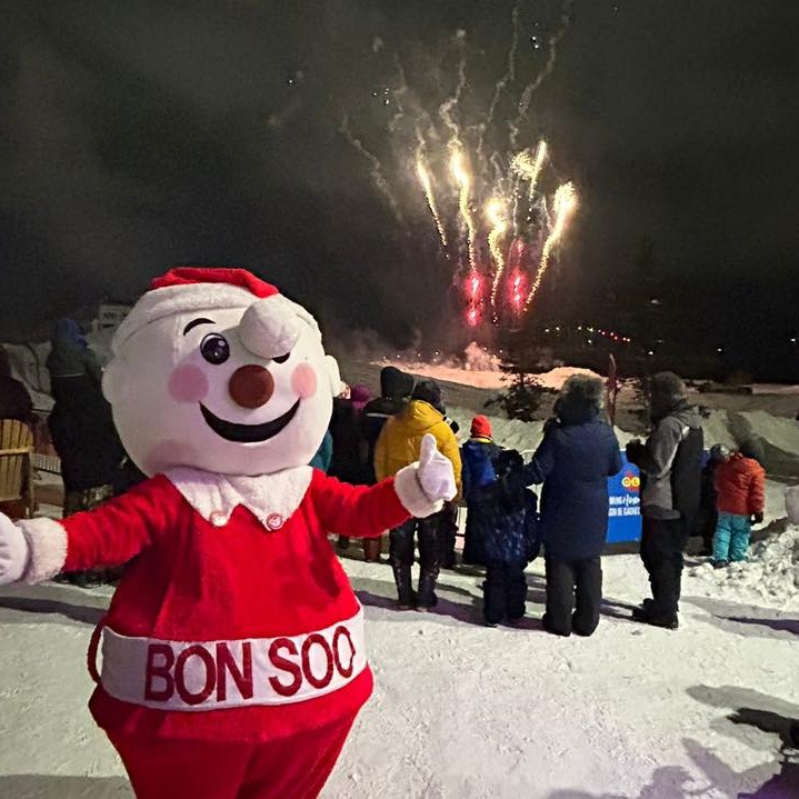 The jolly snowman mascot Mr. Bon Soo gives the thumbs up in front of a crowd of visitors watching an outdoor fireworks display on a snowy night in Sault Ste. Marie at the Bon Soo Winter Carnival. 