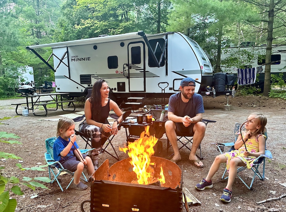 Family of four sitting around campfire with trailer in background.