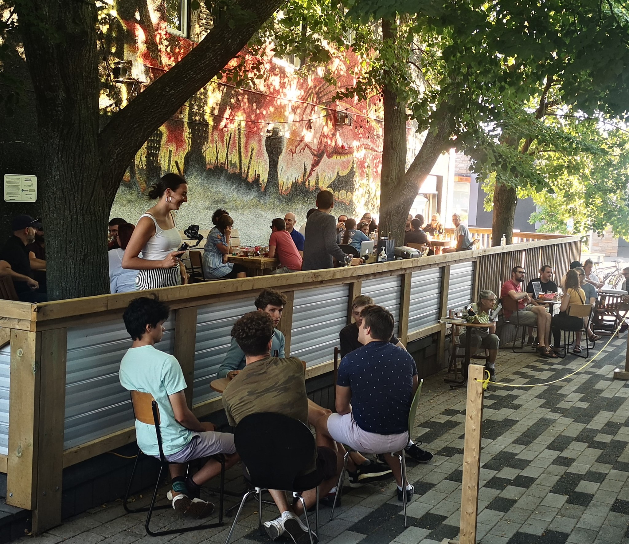People smile and chat on a bustling, tree-shaded streetside patio on a summer day at Outspoken Brewery. 