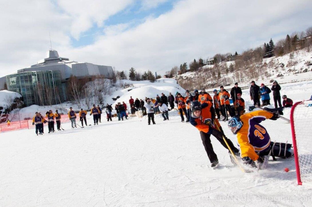 A player shoots on a goal on frozen Ramsey Lake as the goalie dives for the save at the Pond Hockey on the Rock Festival. A crowd of spectators stand down the ice, with the Science North building on the shore. 