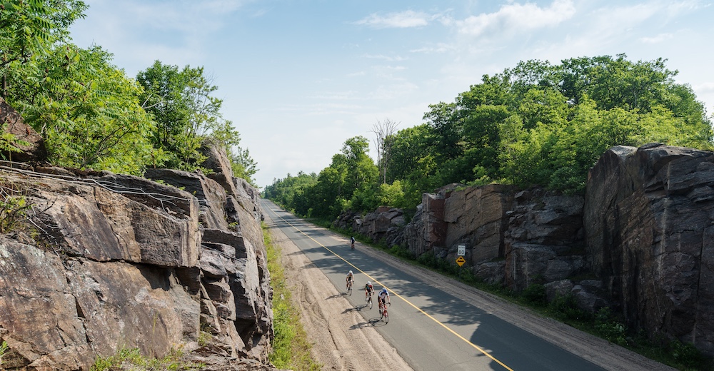 Road cyclists riding single file along road with tall rock cut on either side