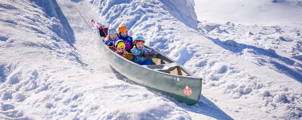 a group of young children smile and laugh as they ride wide-eyed down an ice slide in a metal canoe at the Bon Soo Winter Carnival in Sault Ste. Marie.