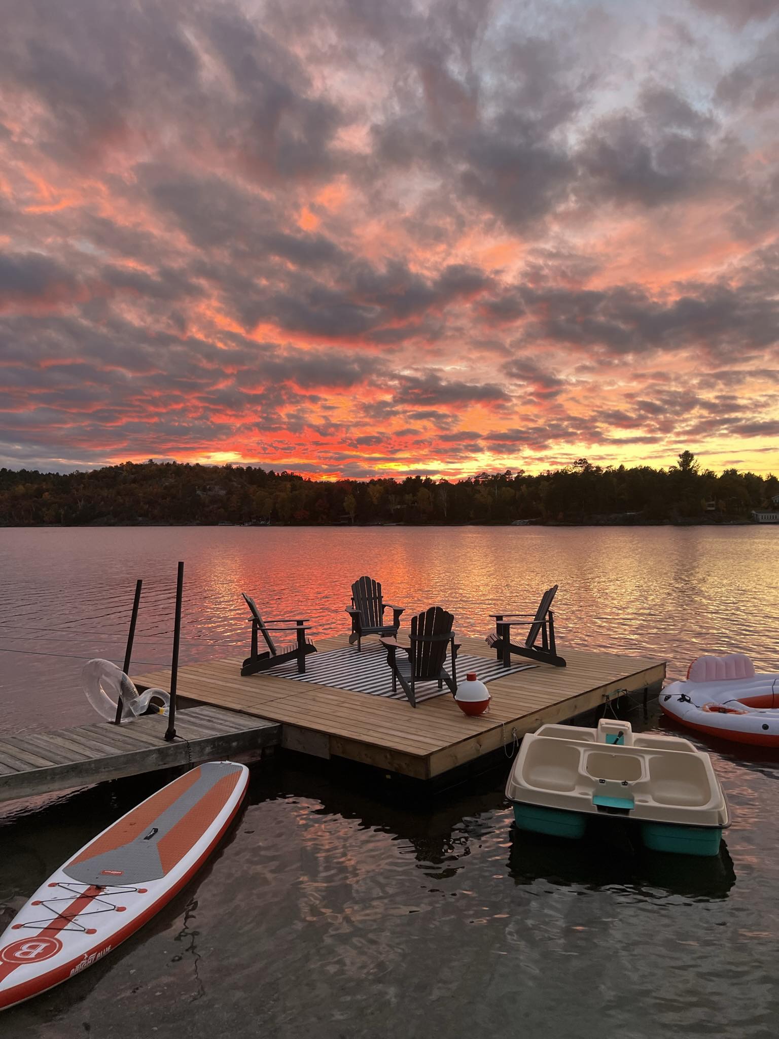 wooden deck chairs arranged in a circle on a wooden dock surrounded by paddleboards and boats under an orange sunset. 