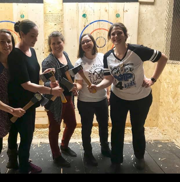 a group of women laugh and hold up their axes together in front of the targets at Northern Axeperts in Sudbury.