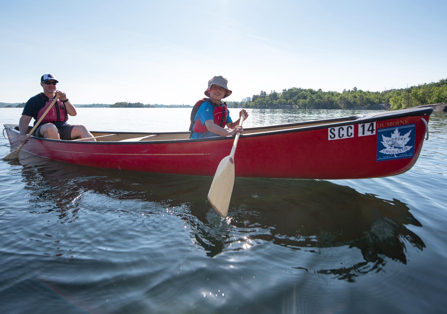 Two paddlers move their canoe labelled "Sudbury Canoe Club" across the glassy surface of a lake on a sunny summer day.