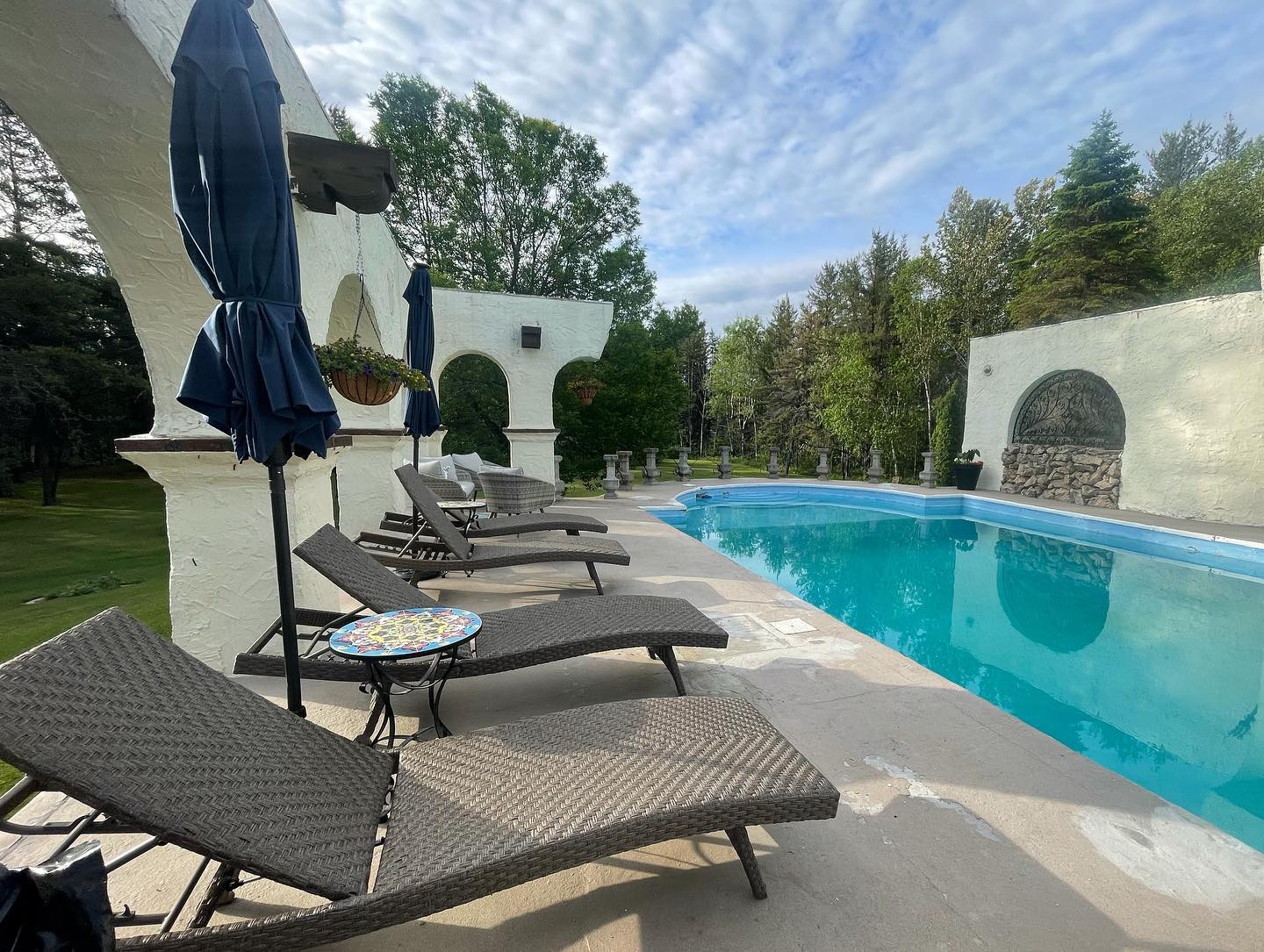a row of lounge chairs alongside a turquoise swimming pool surrounded by green lawns and trees under a summer sky at The Hacienda Bed and Breakfast near Sudbury. 