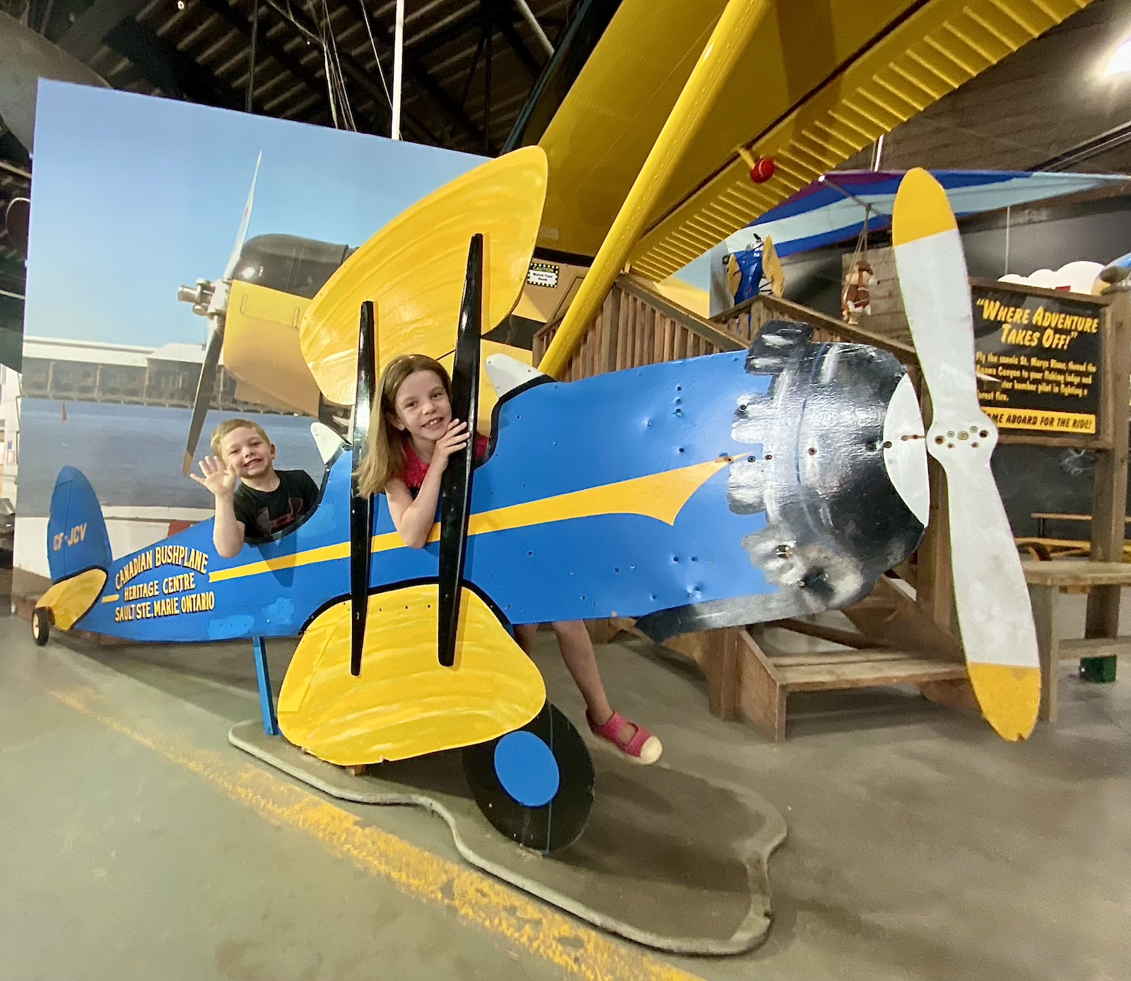 two kids smile and wave from the seats of a large model bushplane at the Canadian Bushplane Heritage Centre.