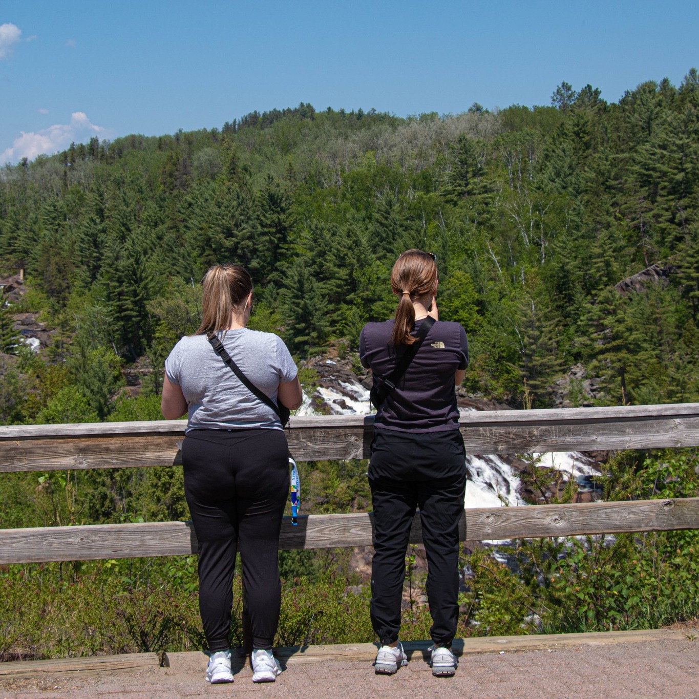 two women shoot pictures of Onaping Falls over a wooden rail along a hiking trail on a sunny summer day. 