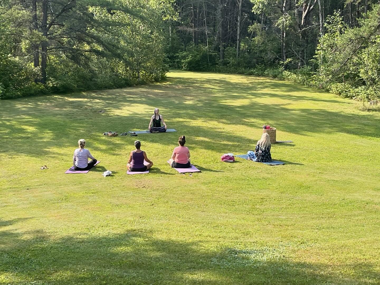 a group of ladies sit in a circle on their yoga mats on a rolling green lawn surrounded by green trees on a sunny day at The Hacienda Bed and Breakfast.
