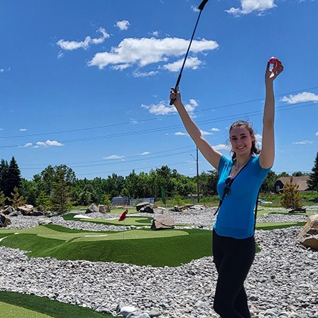 a woman smiles as she poses with her golf club and ball held in the air on a mini putt course in Sudbury, Ontario.