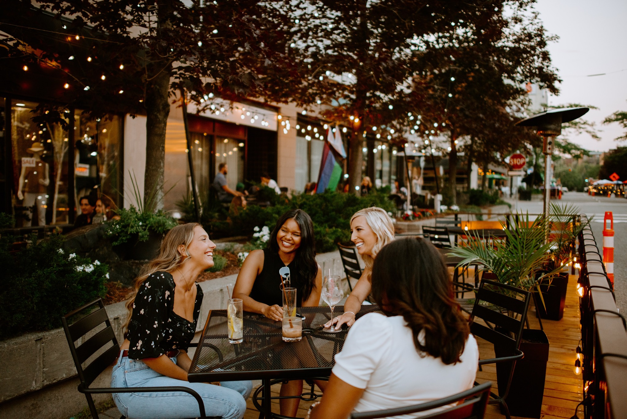 a group of ladies laugh and chat around a table over drinks on a patio in the summer. The green trees around the patio are lit up by twinkle lights. 