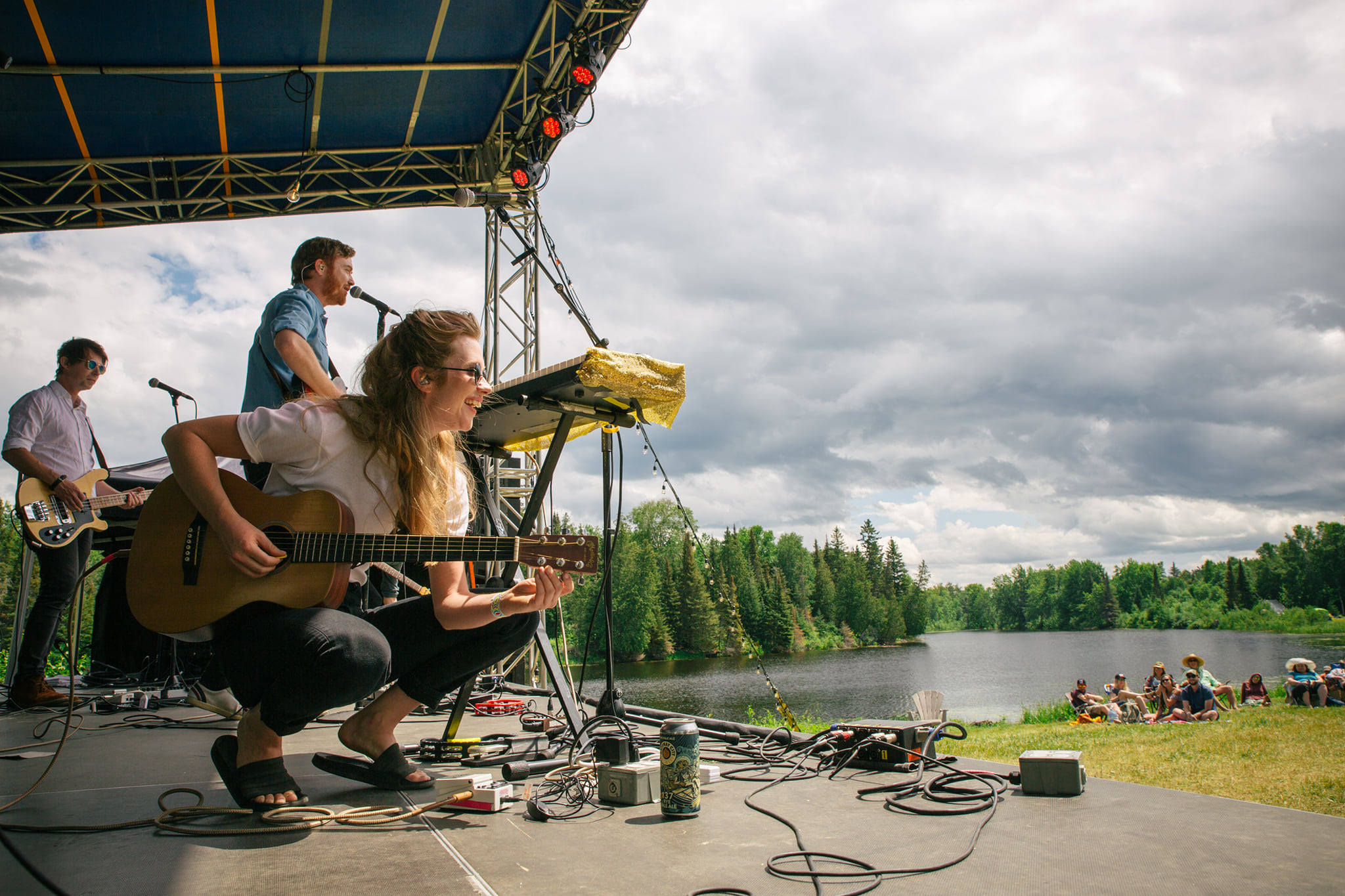 a smiling guitar player crouches onn stays as she sings to the crowd on the green grass next to a shining river at the River & Sky music festival.