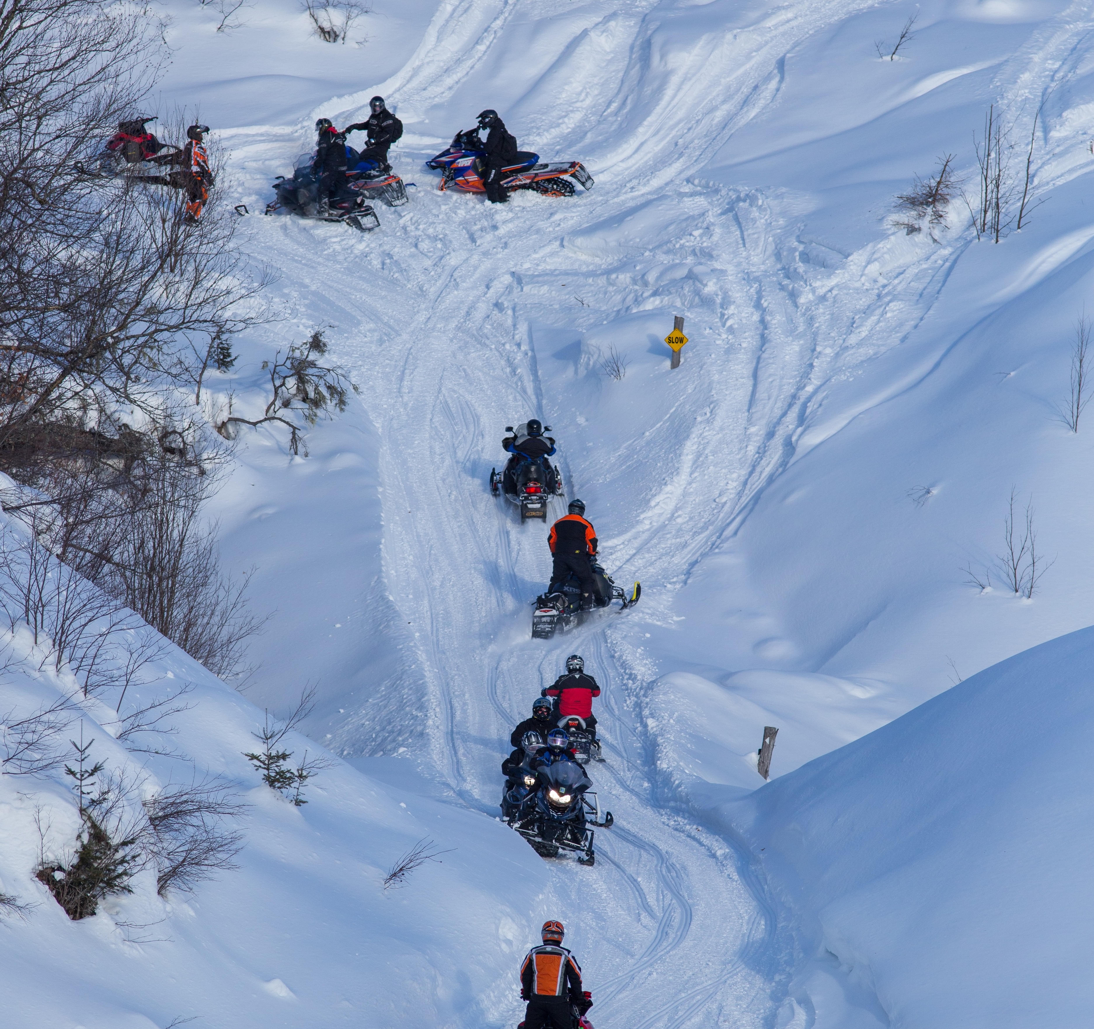 many snowmobilers at once ride through a gully filled with deep powder snow.