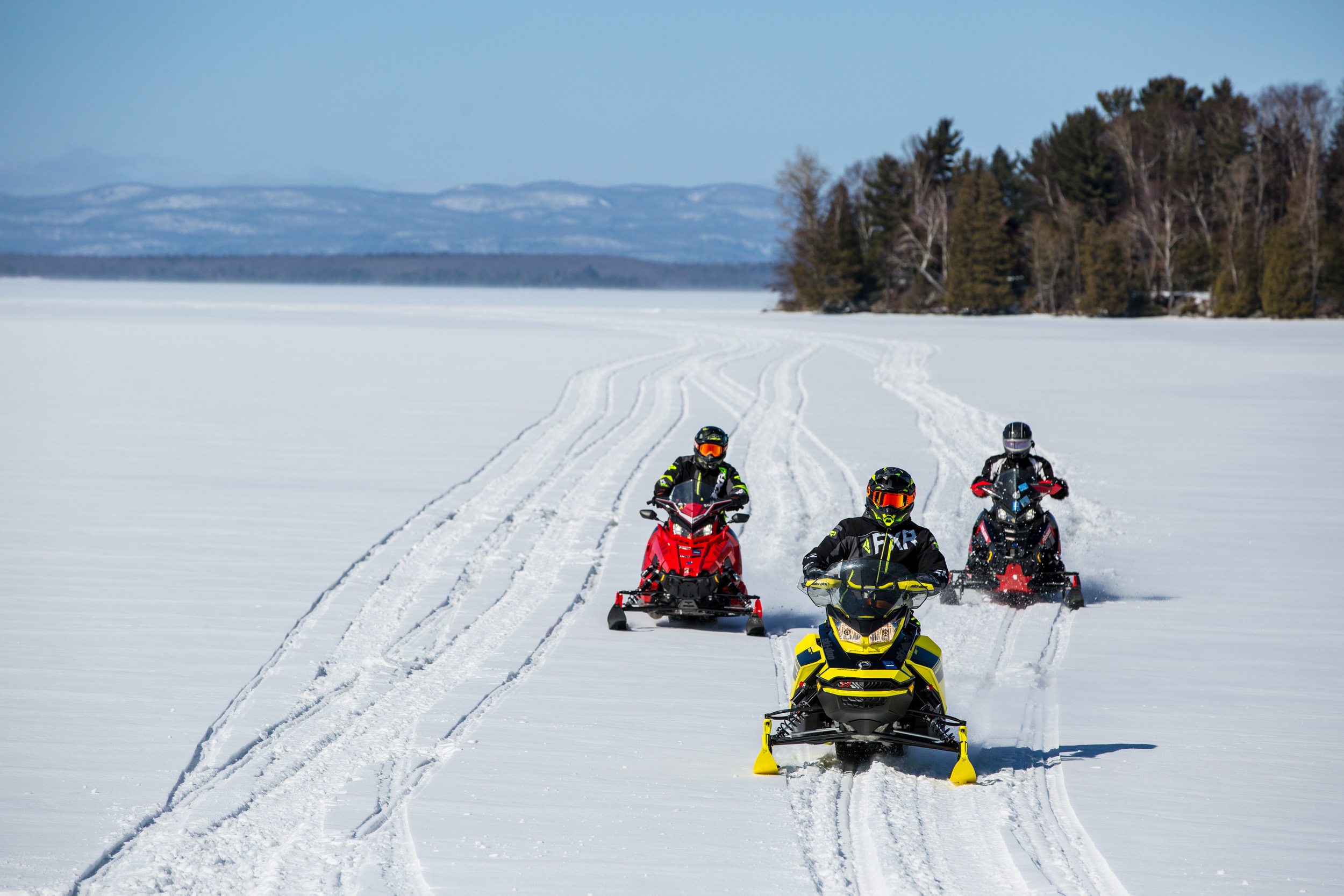 3 snowmobilers ride across a frozen, snow-covered lake surrounded by forest on a sunny winter day. 