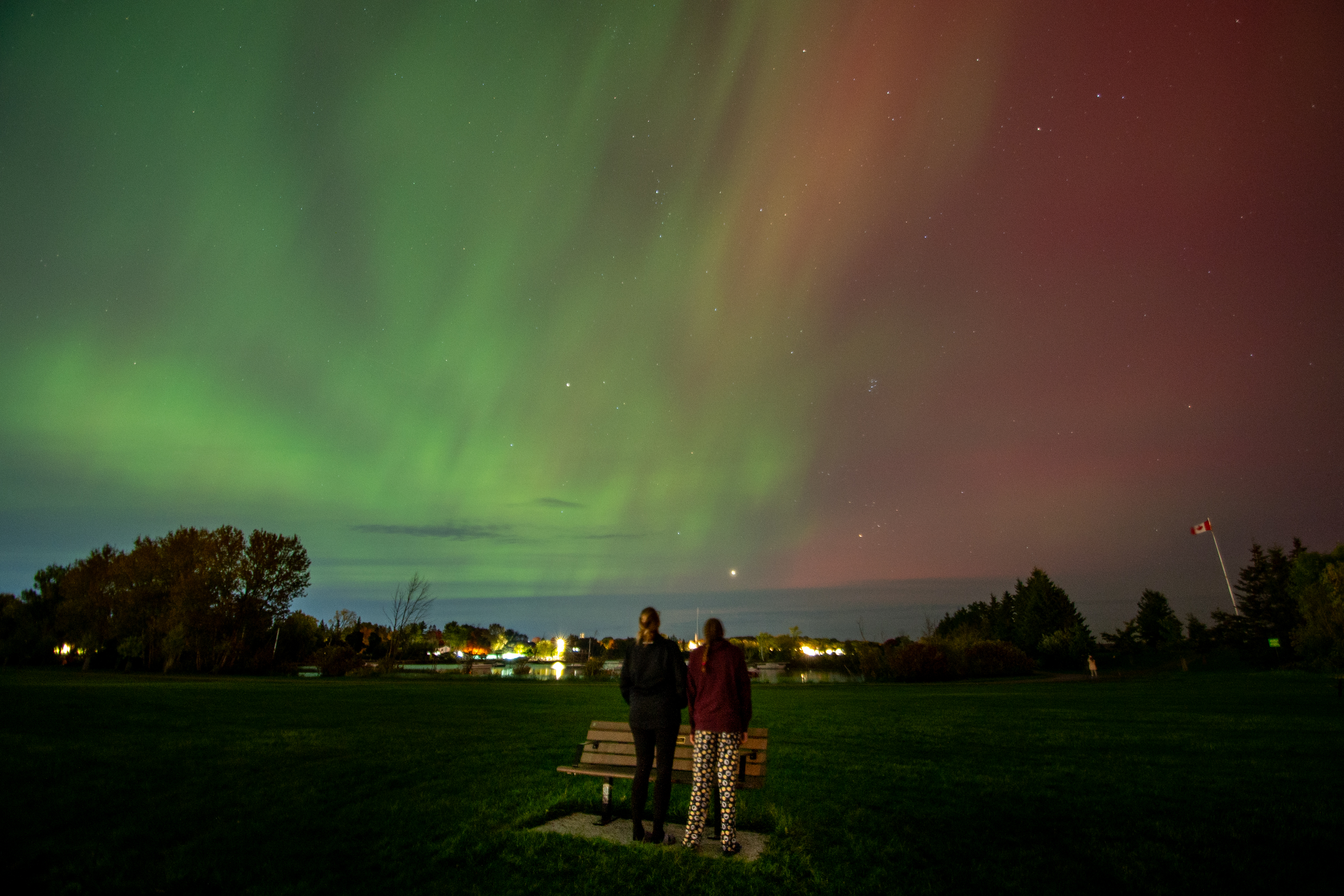 two people stand close together looking up at the swirling green, red and yellow aurora borealis that fills the sky over Lake Superior in Sault Ste. Marie. 