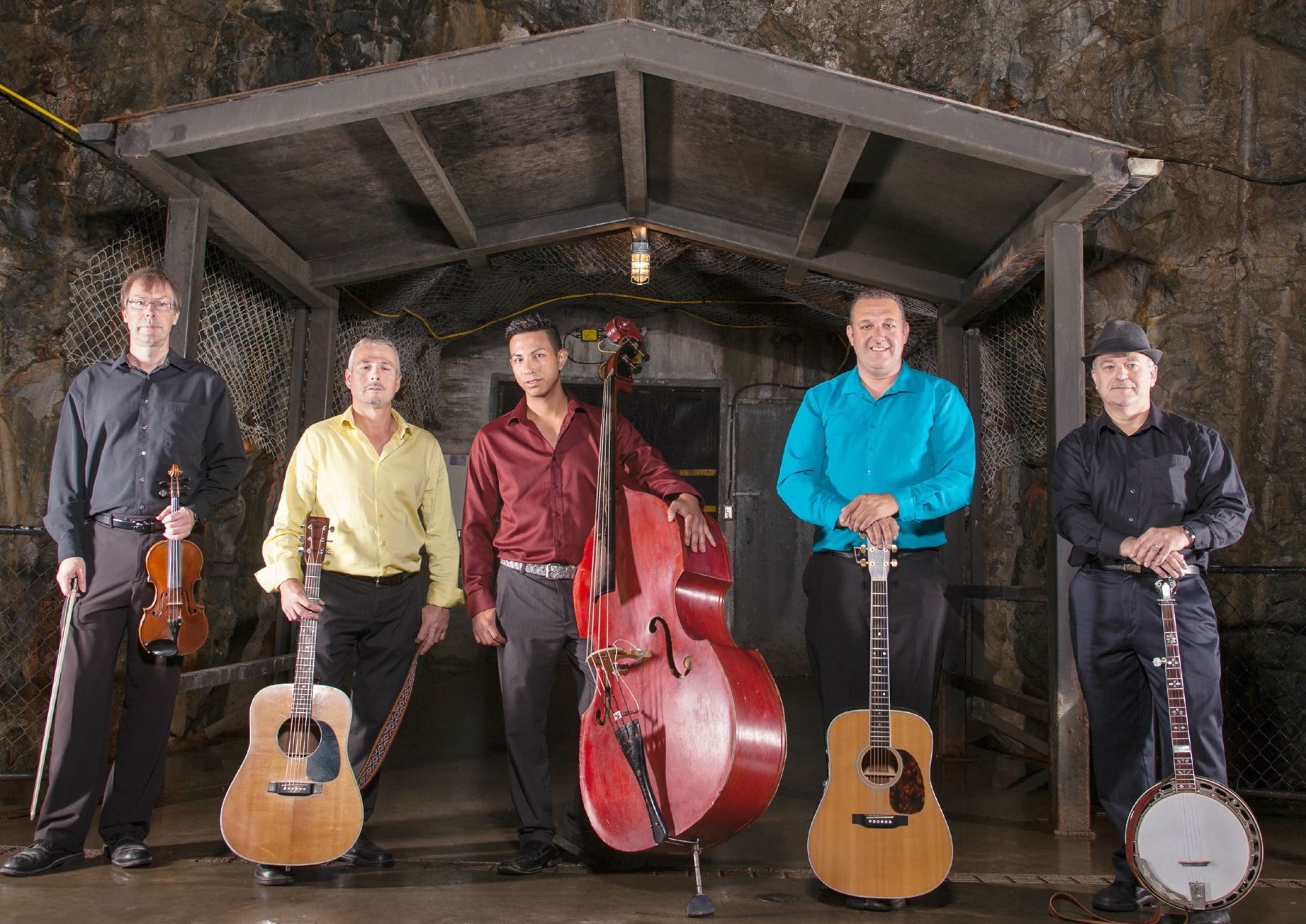 Canucky Bluegrass Boys, a smartly dressed group of musicians standing with their string instruments at the rocky opening of a mine.  