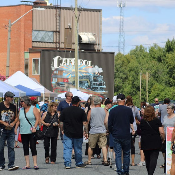 People mill about main street next to a series of makret tents on a sunny summer day. A mural with the word "Capreol" is painted on the side of a brick building in the background.  