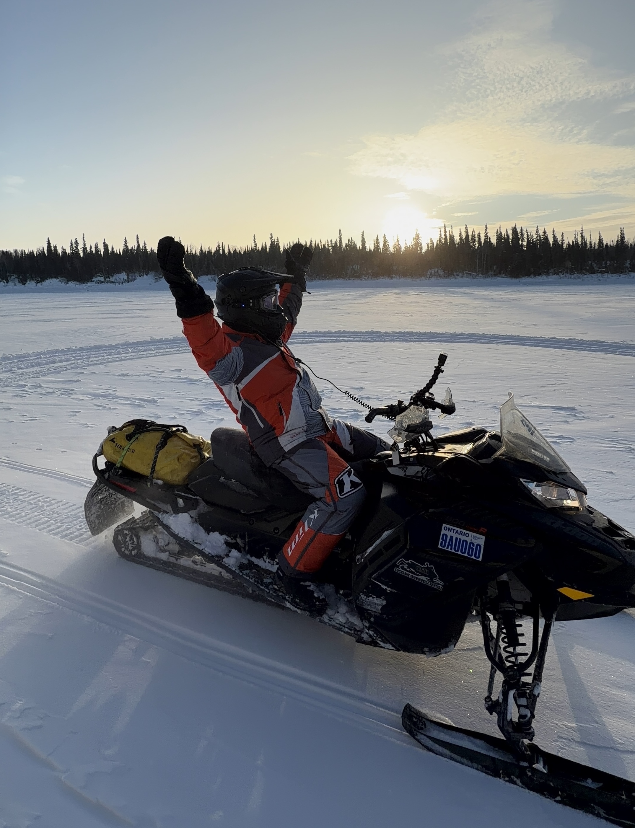 Eric on his snowmobile with arms raised, looking at the vast frozen strectch of snow around him and the setting sun. 
